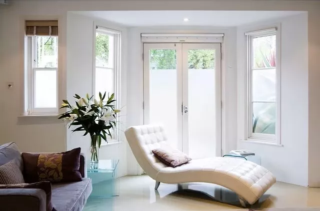 Bright modern living room with white chaise lounge, glass side table, lilies, and French doors with frosted glass.