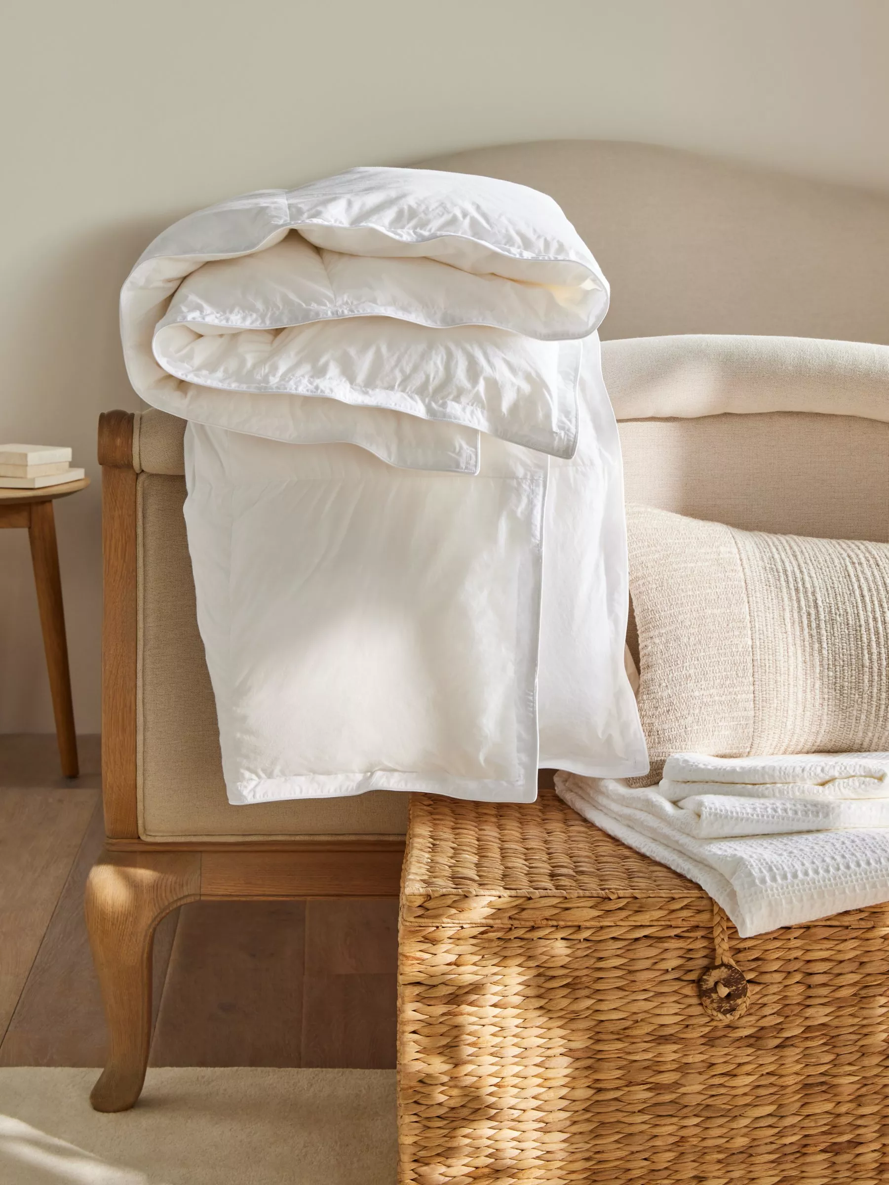 A neatly folded white duvet draped over a cream upholstered bench, with a wicker basket nearby.