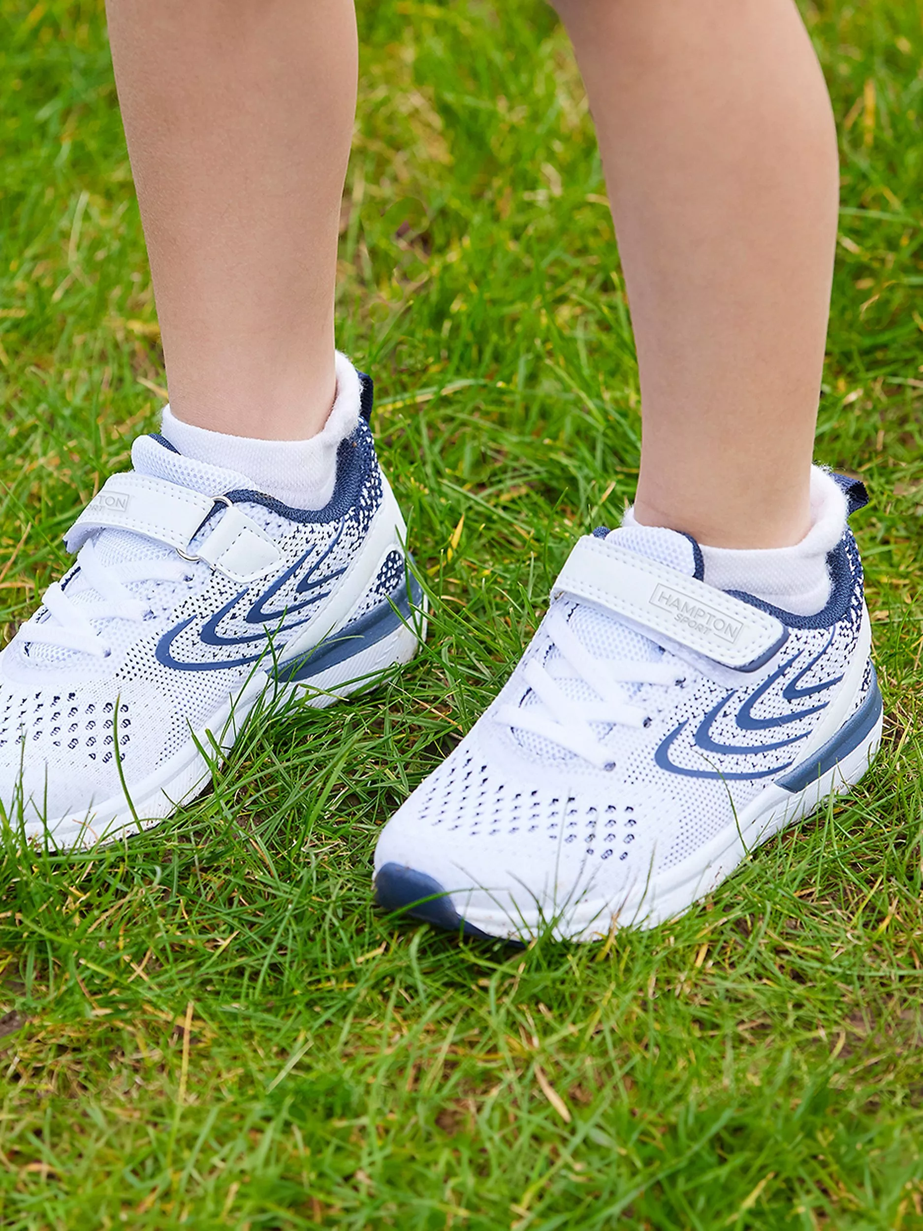 Close-up of child wearing white and navy Velcro trainers and socks, standing on green grass outdoors