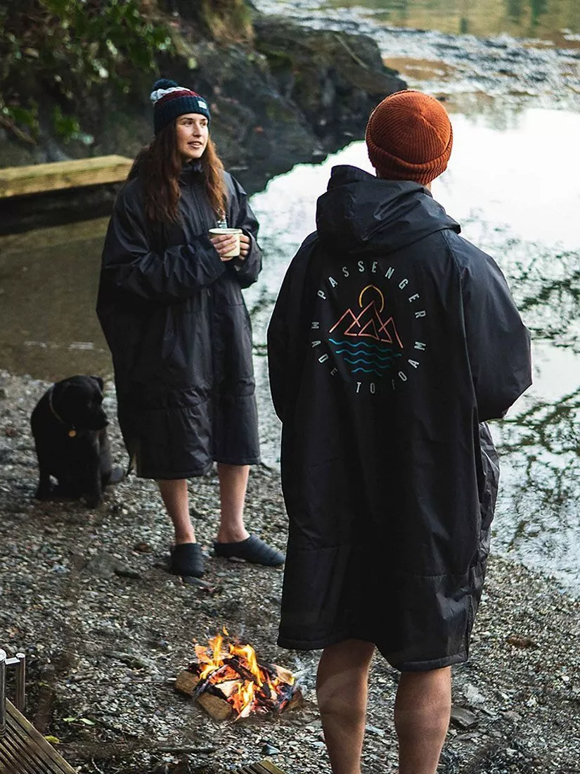 Two campers in waterproof changing robes by a lakeside campfire, with a dog nearby, outdoor adventure scene