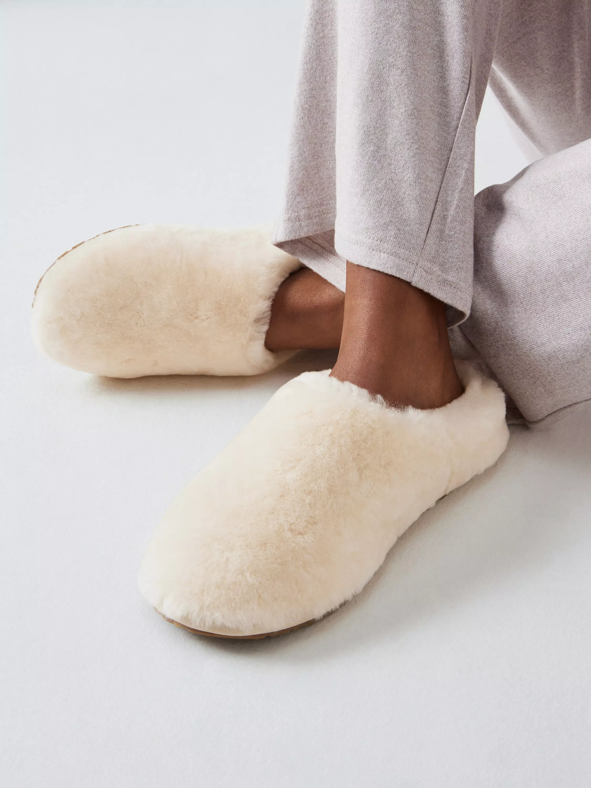 Close-up of cosy cream faux fur slippers worn with grey loungewear trousers on a light studio background