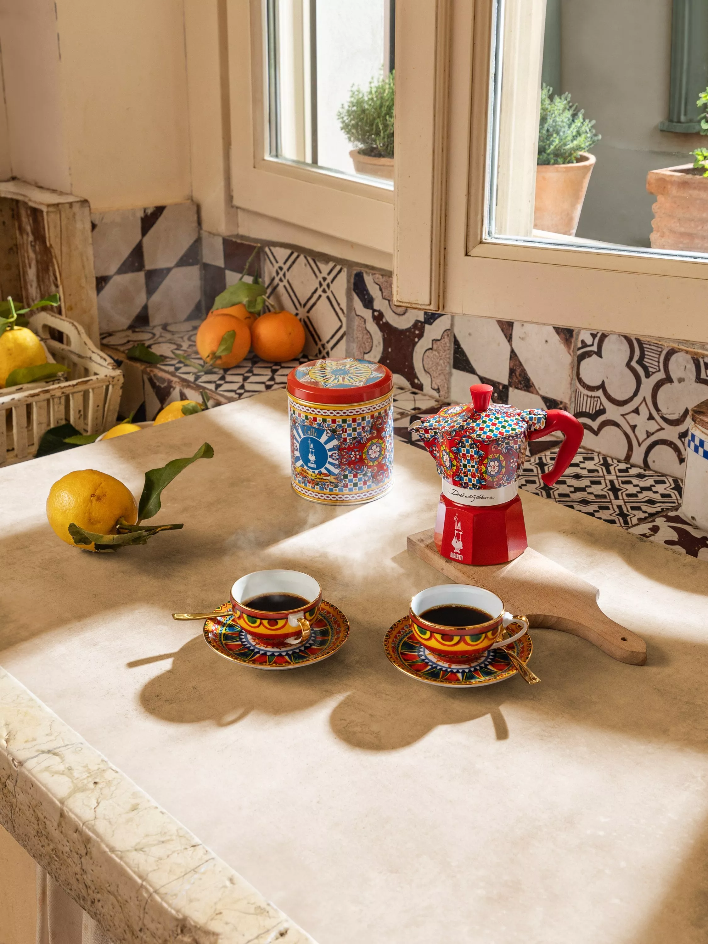 Colourful coffee setup with two steaming cups on a tiled counter, surrounded by oranges and lemons near a window.