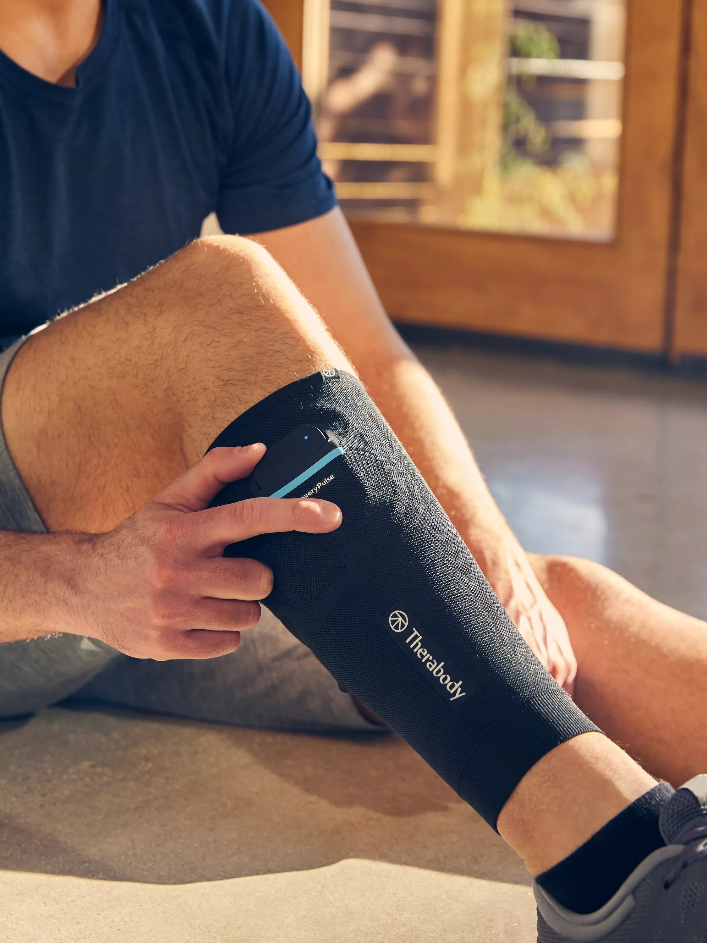 Athlete using Therabody calf compression sleeve with control unit, seated on gym floor in sunlight.