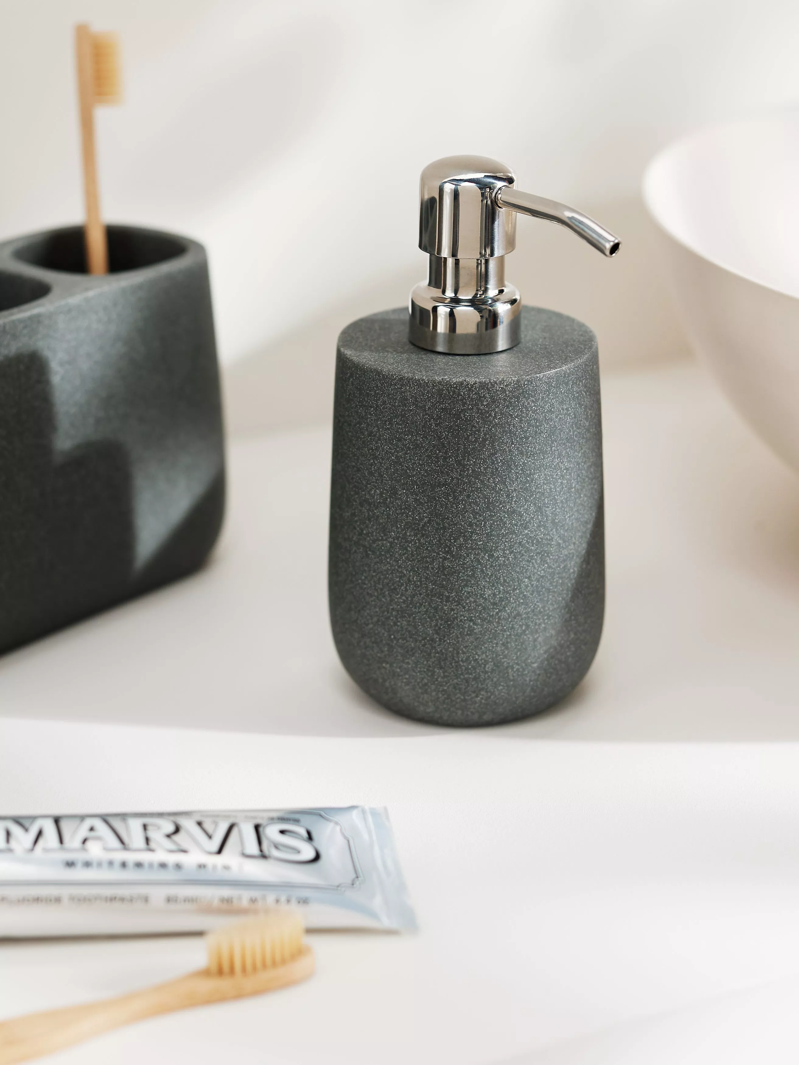 Grey stone soap dispenser with chrome pump on white bathroom counter, bamboo toothbrush and toothpaste tube nearby