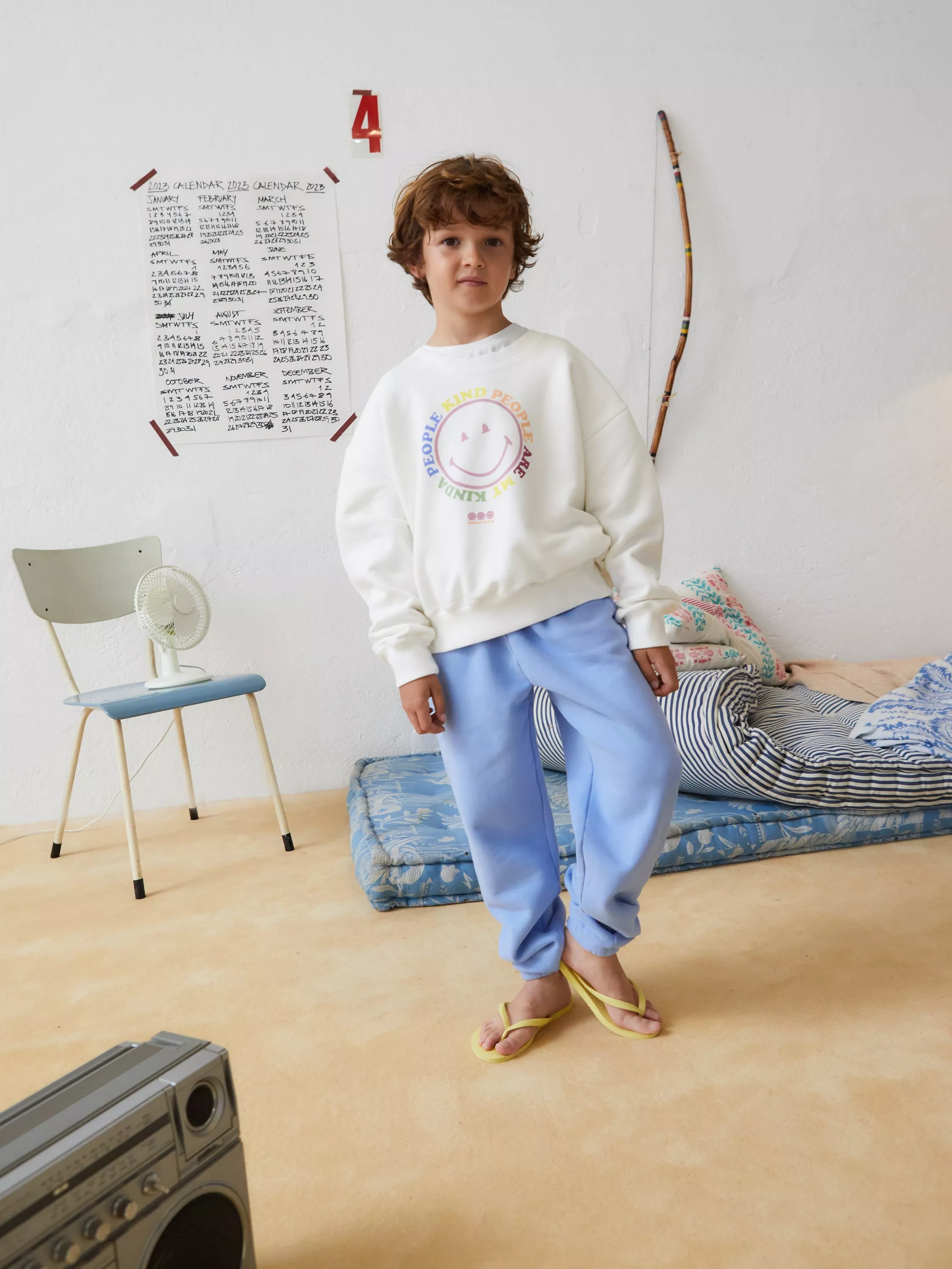 Child modelling white graphic sweatshirt and blue joggers in bedroom with floor mattress and wall calendar.