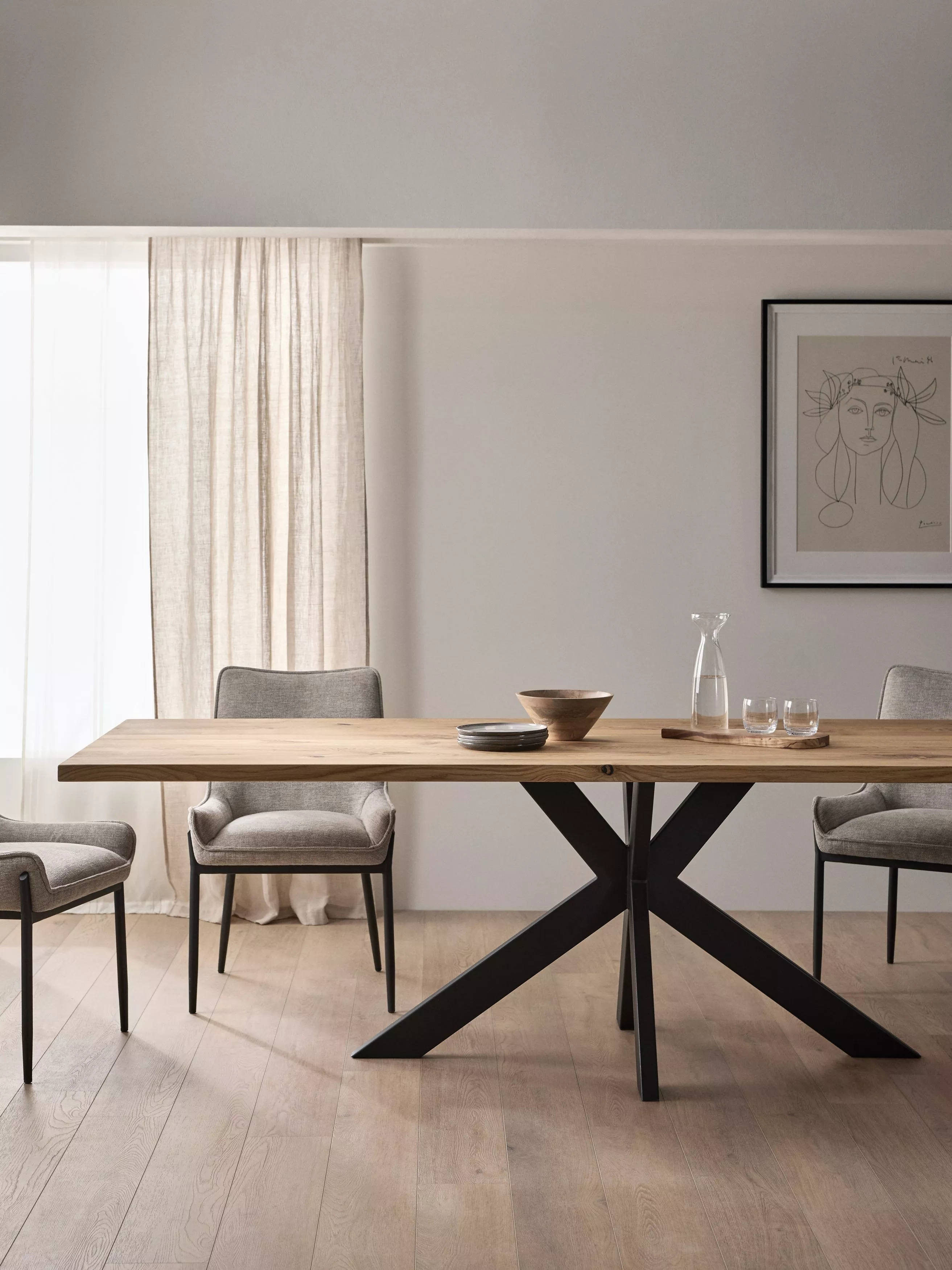 Modern dining room with wooden table, grey chairs, and artwork on the wall. Natural light filters through curtains.