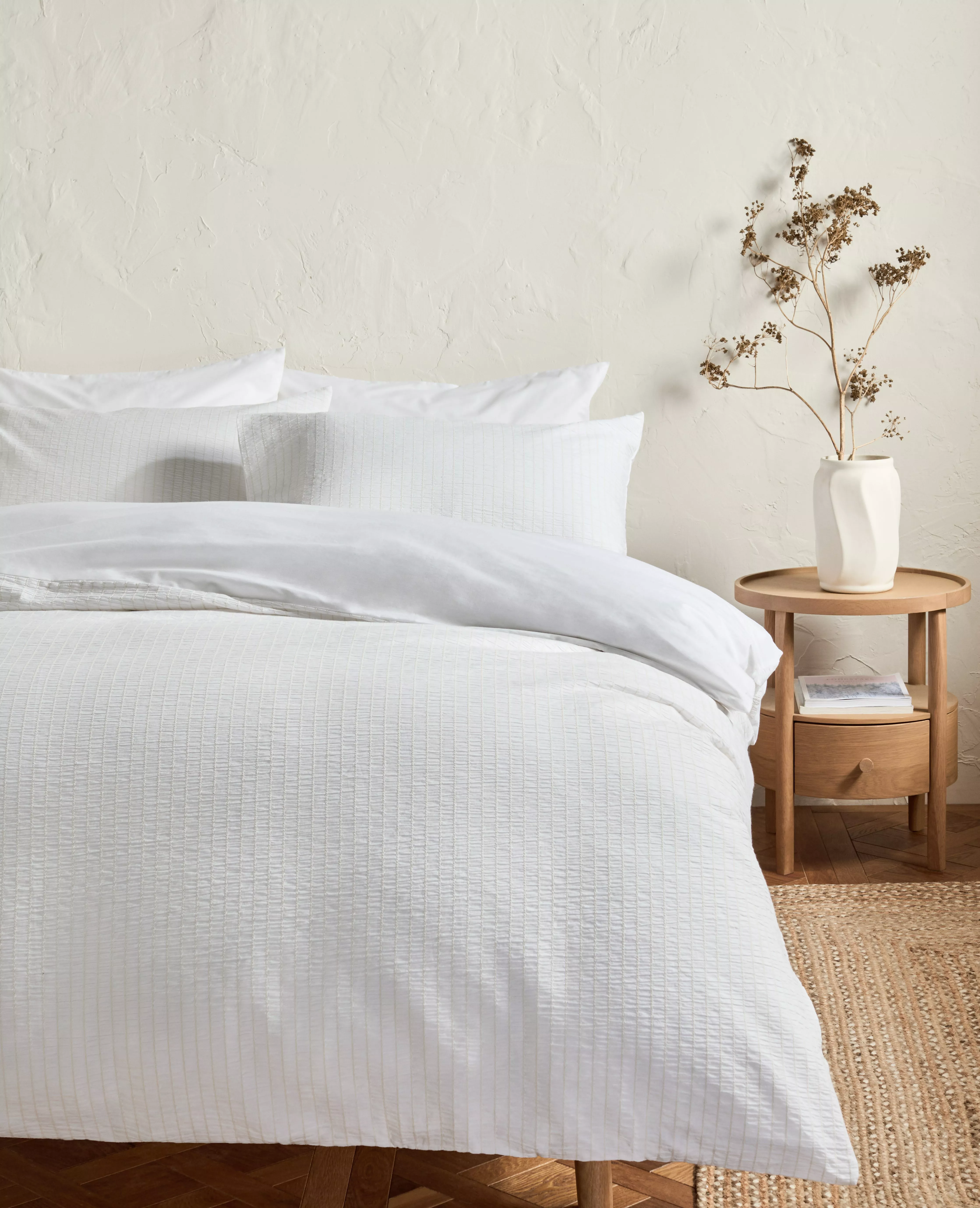 Minimalist bedroom with white textured duvet cover and pillows, beside wooden bedside table and vase