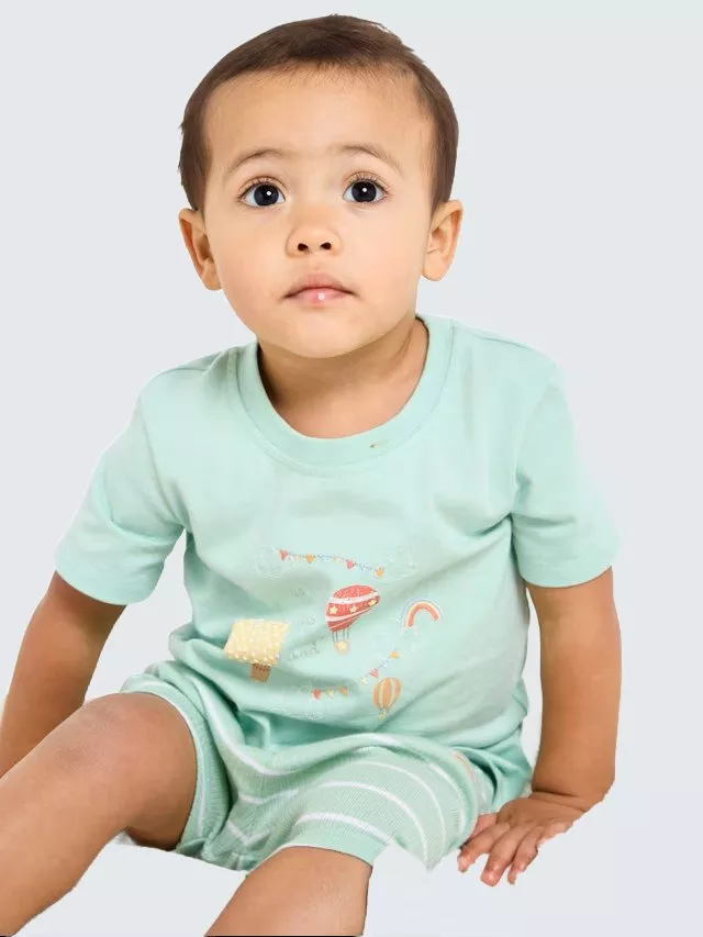 A toddler in a mint green outfit with playful graphics sits against a neutral background, looking upwards.