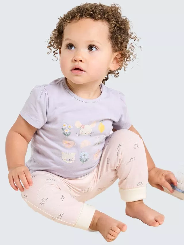Toddler with curly hair wearing a lilac top and patterned trousers, sitting and looking upwards.