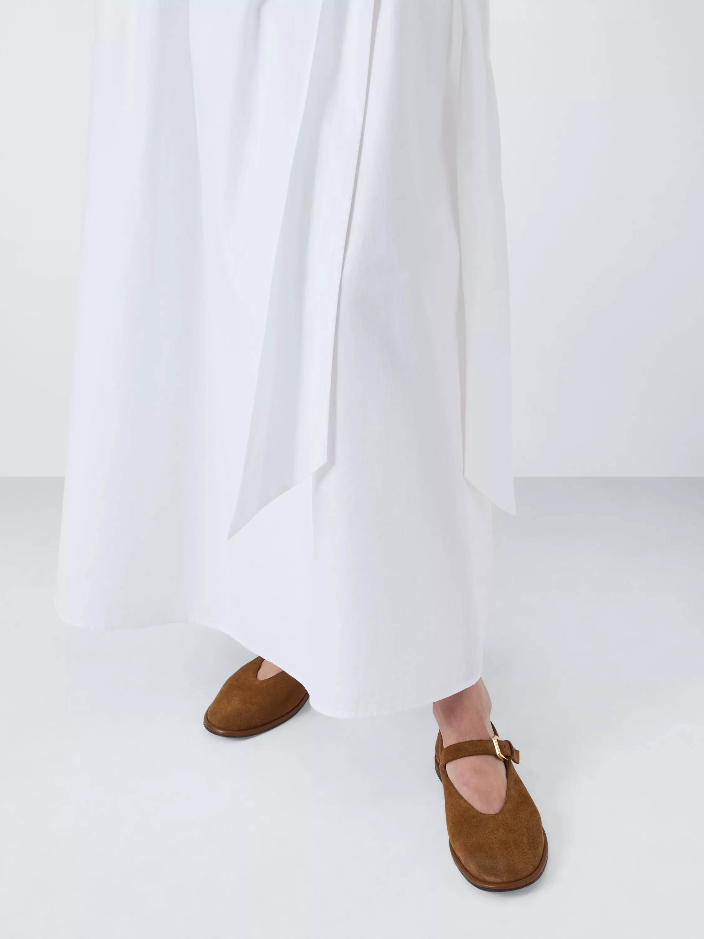 Close-up of white maxi dress hem with brown suede Mary Jane flats, minimalist studio fashion shot