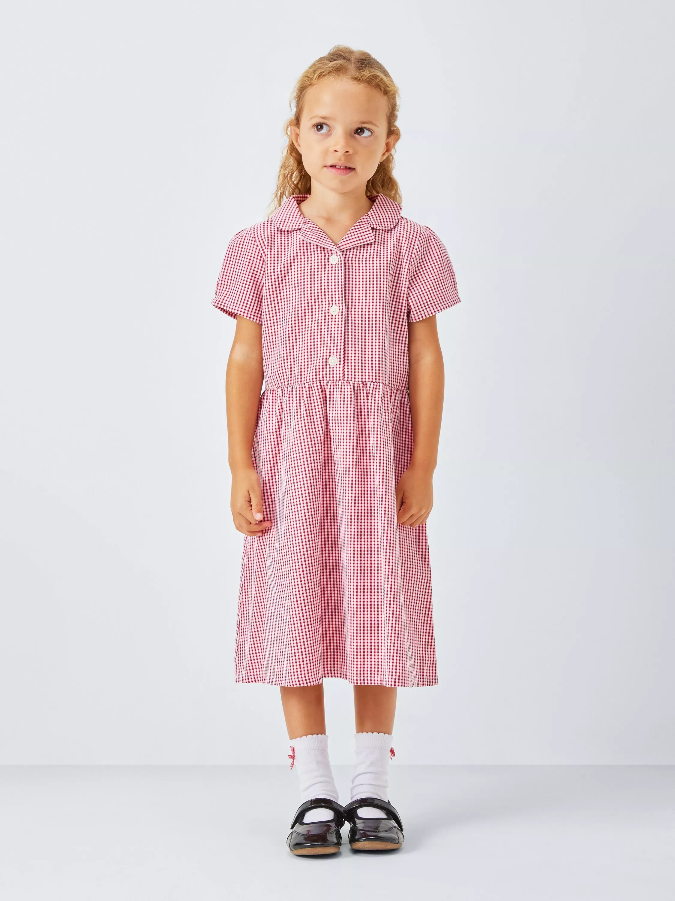 Young girl in red gingham school dress with white socks and black shoes, standing against plain backdrop
