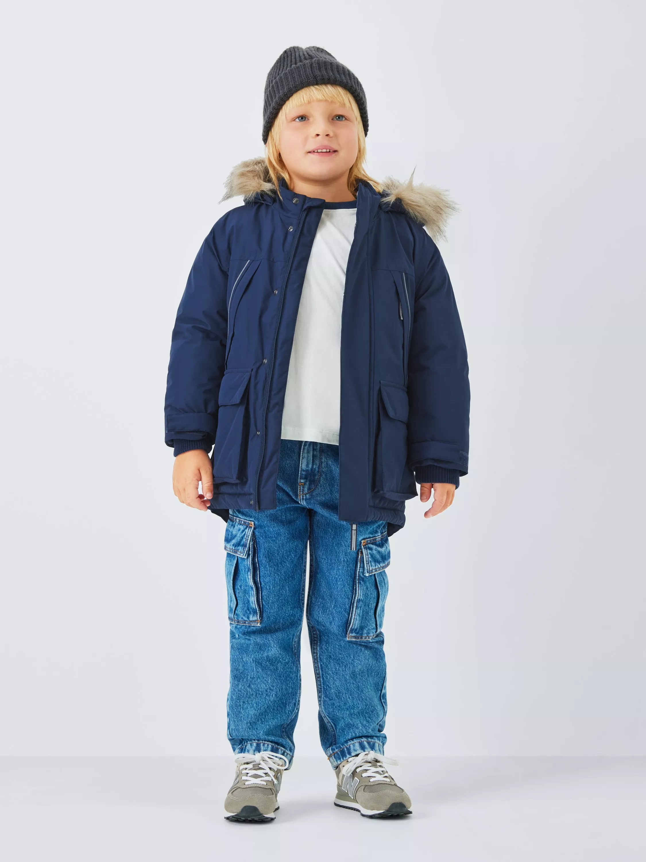 A boy wearing a navy blue parka with a faux-fur hood, a grey beanie, blue cargo jeans, and trainers
