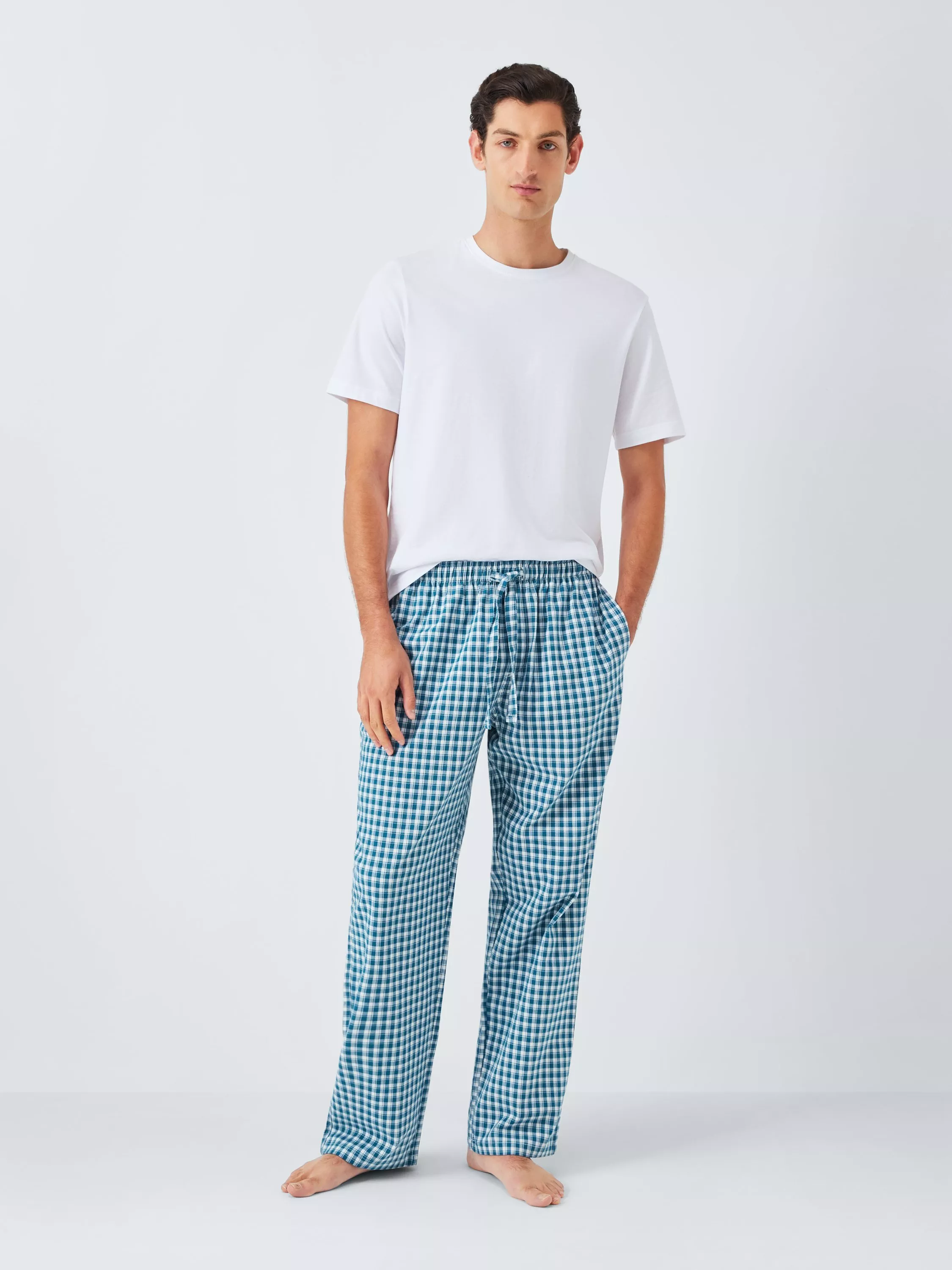 Male model wearing a white crew-neck T-shirt and blue gingham pyjama trousers, standing barefoot in studio.