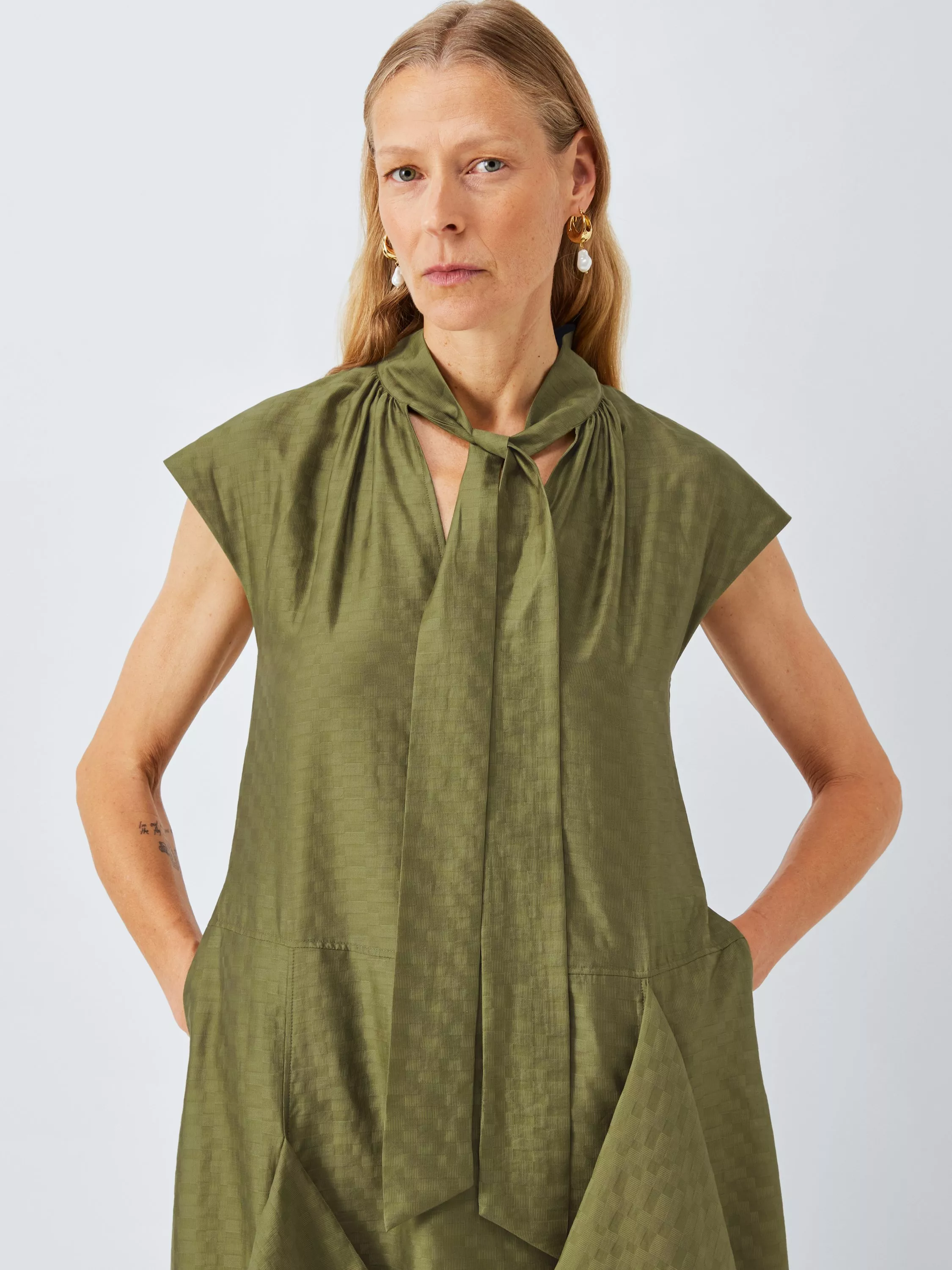 Studio portrait of a woman wearing an olive green tie-neck dress with cap sleeves and pockets on a white backdrop