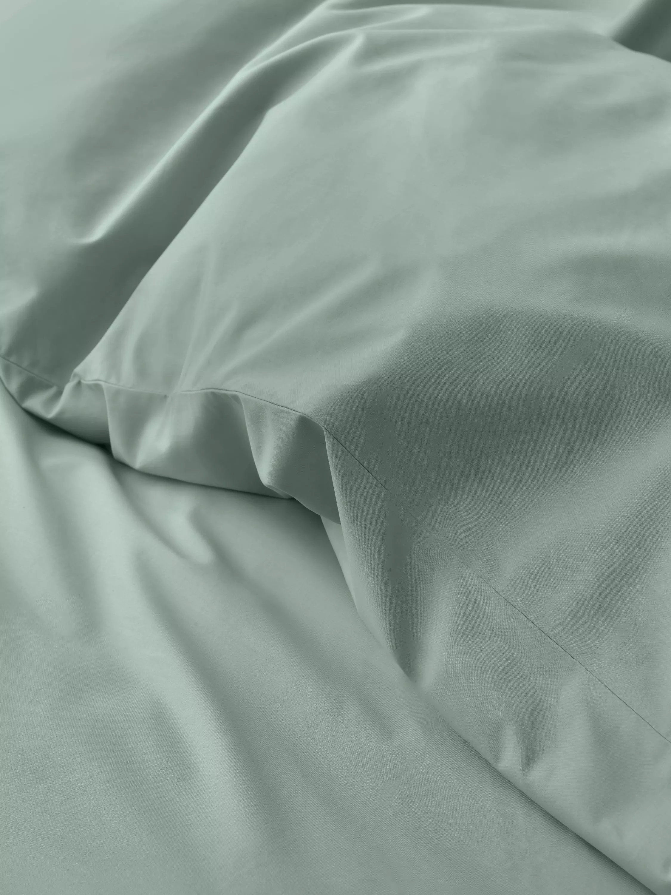 Close-up of sage green pillowcase and bed sheet set with smooth cotton fabric and crisp stitched seams