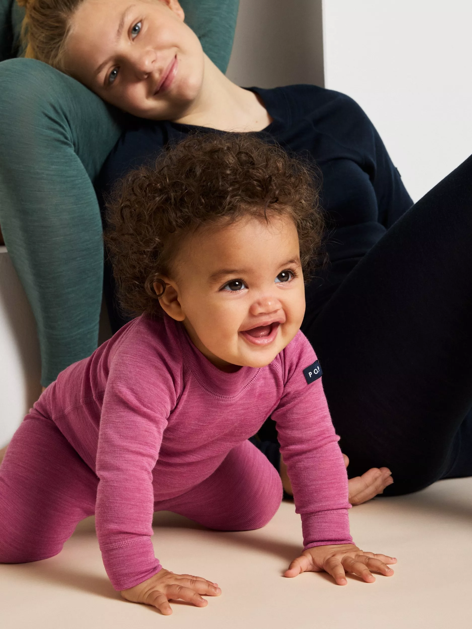 Baby in pink outfit crawls on cream floor, smiling. Person in background leans on knee, wearing dark clothing.