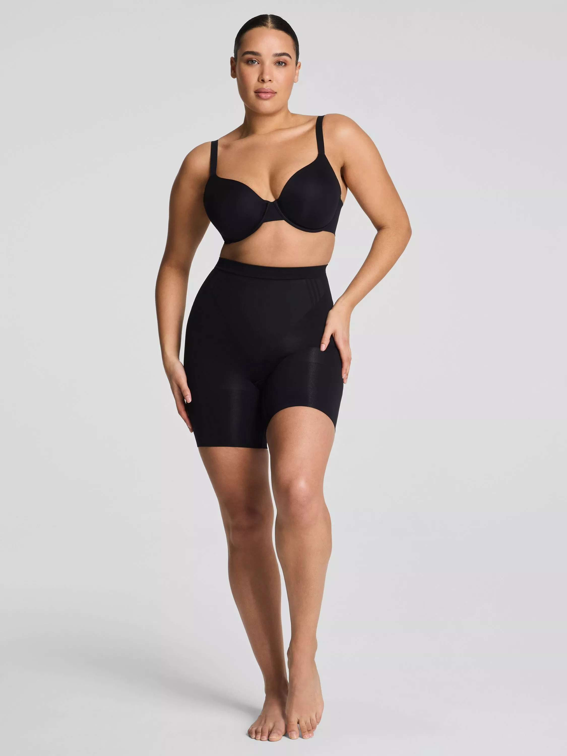 Plus-size model wearing black underwired bra and high-waist shapewear shorts on a light grey studio background