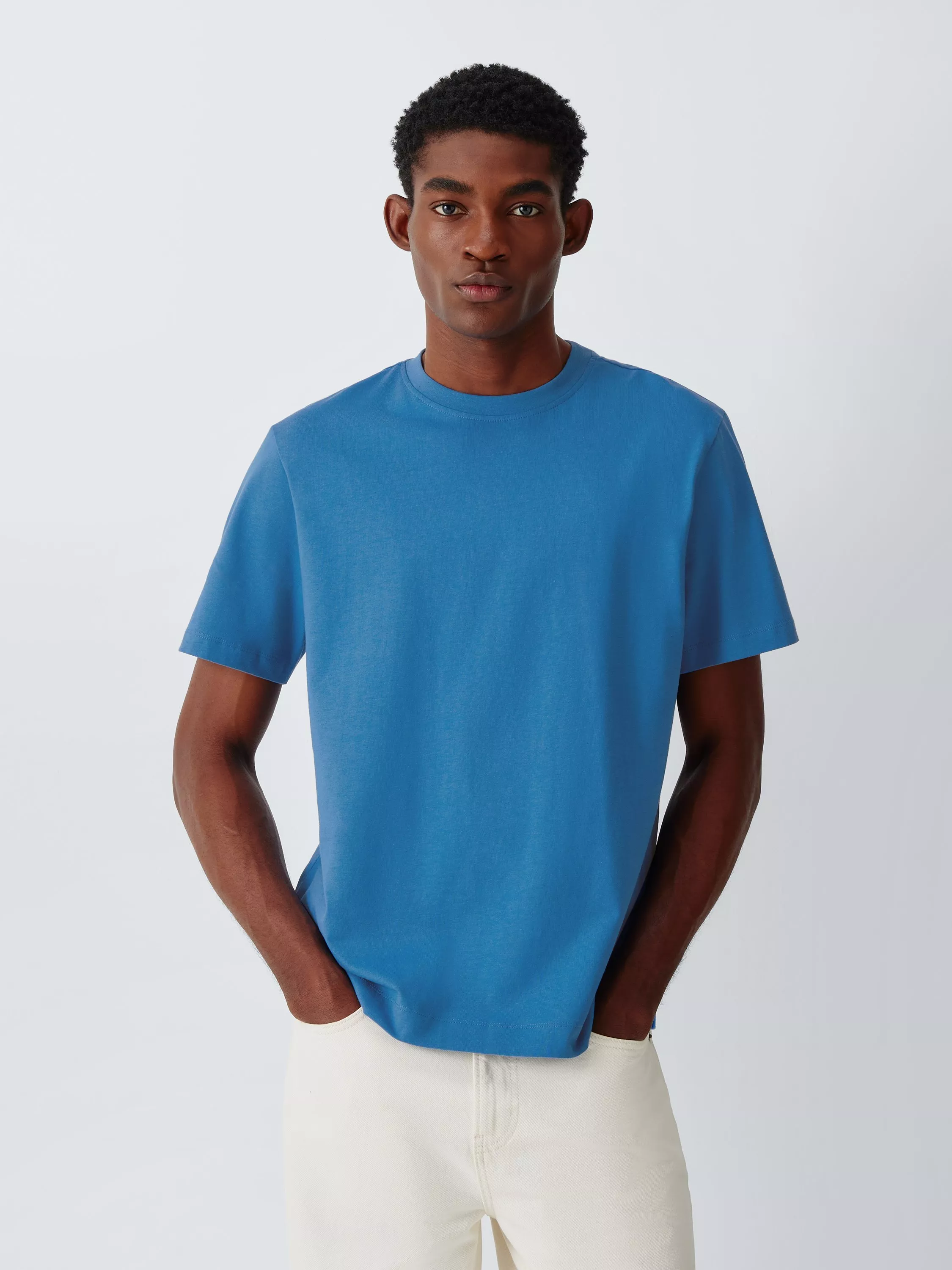Studio portrait of a man wearing a blue crew neck T-shirt and white trousers, hands in pockets on grey background