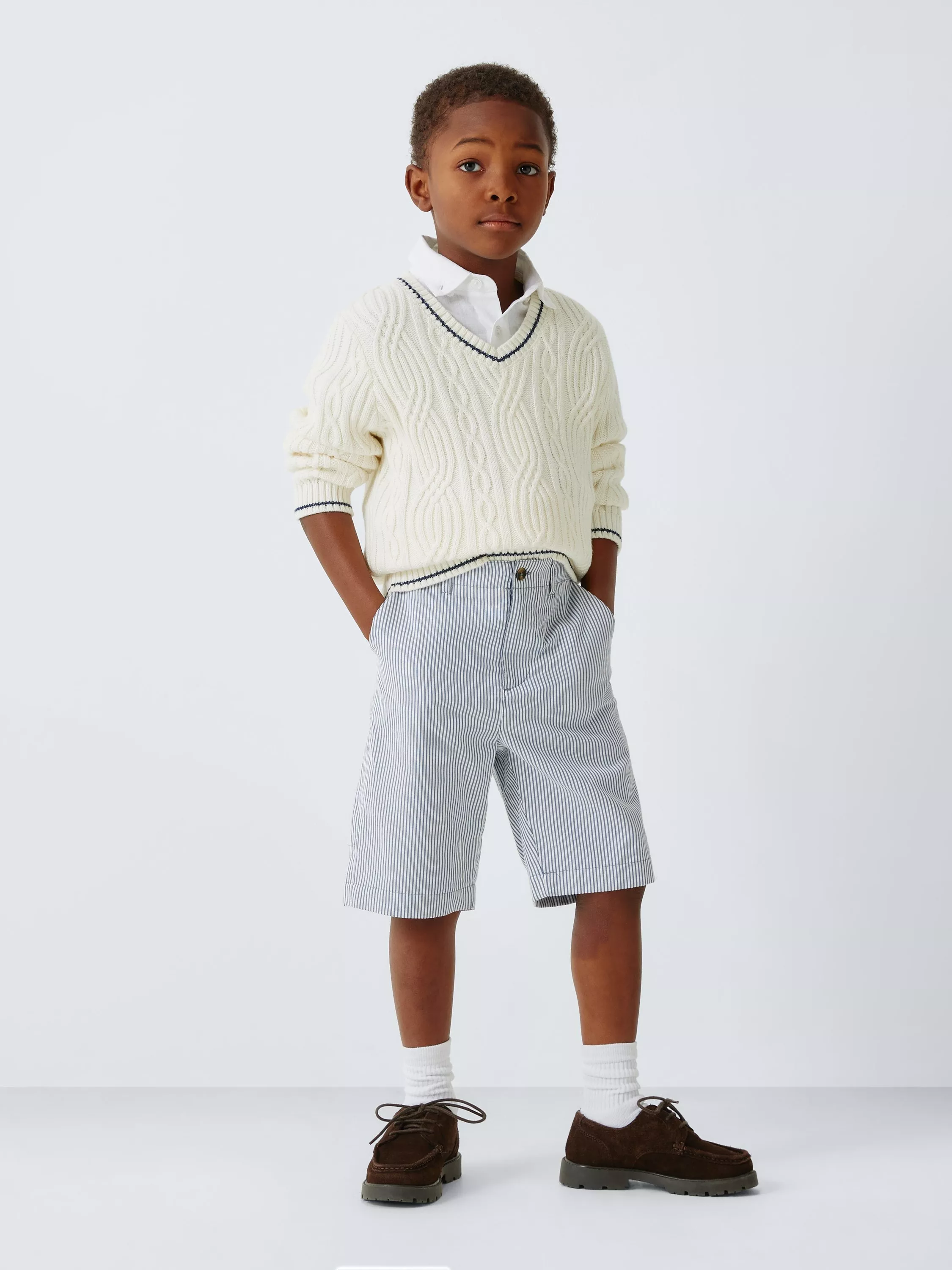 A child in a cream cable-knit jumper and striped shorts stands confidently, hands in pockets.