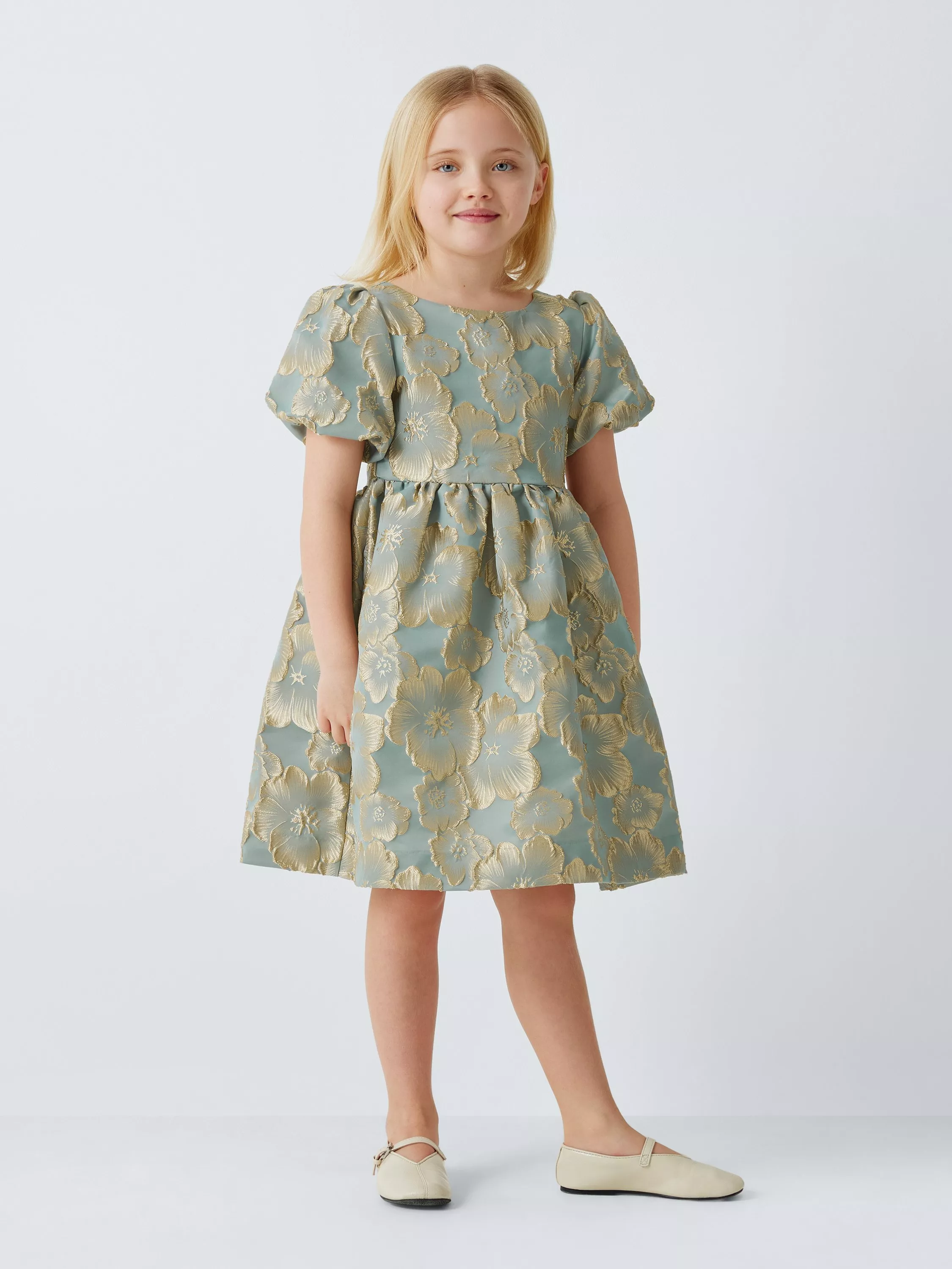 Young girl in a floral dress with puffed sleeves, featuring gold patterns on a light blue background, and beige shoes.
