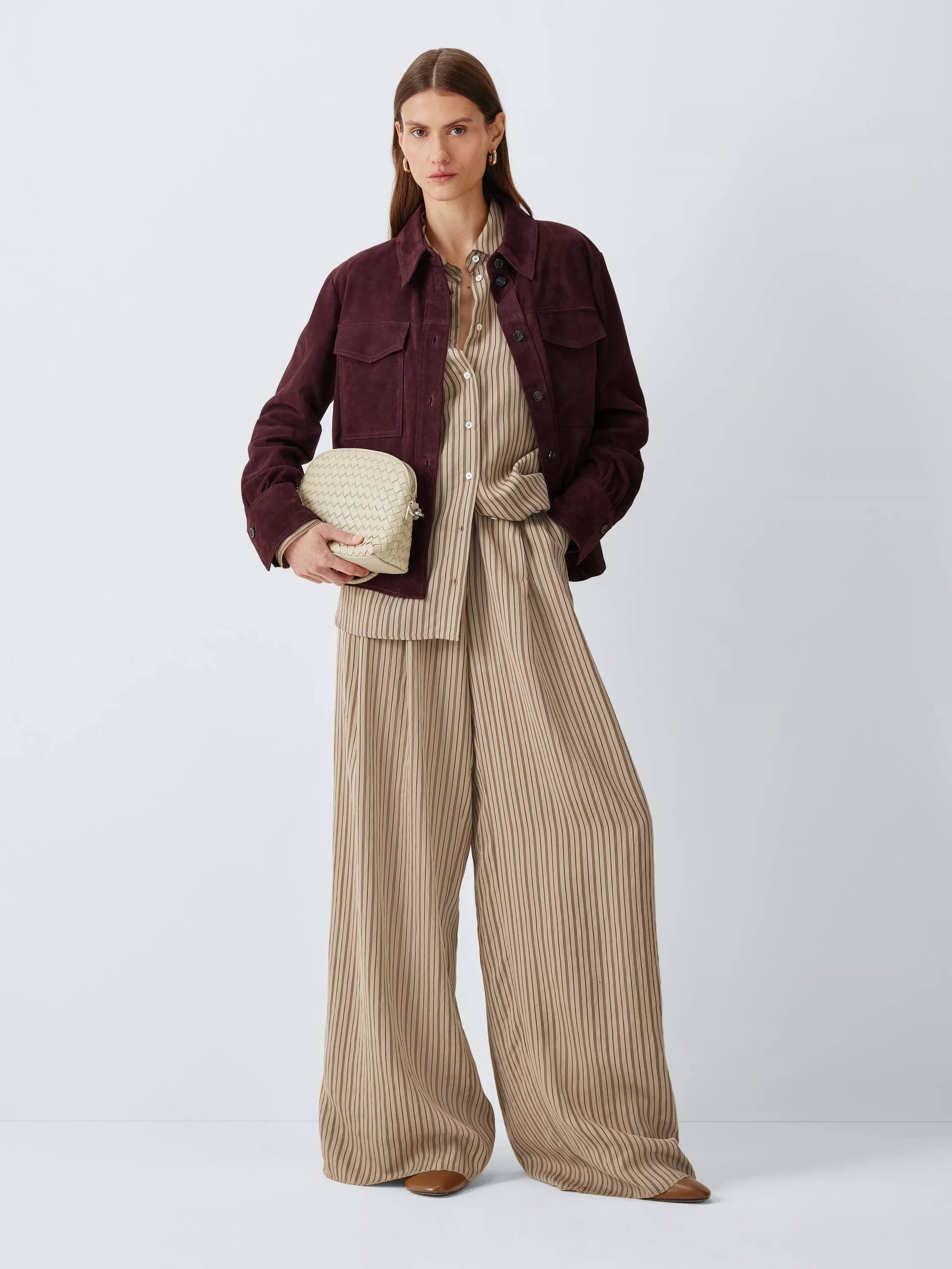 Woman in a burgundy jacket over a striped beige co-ord, holding a woven clutch, standing against a neutral background.