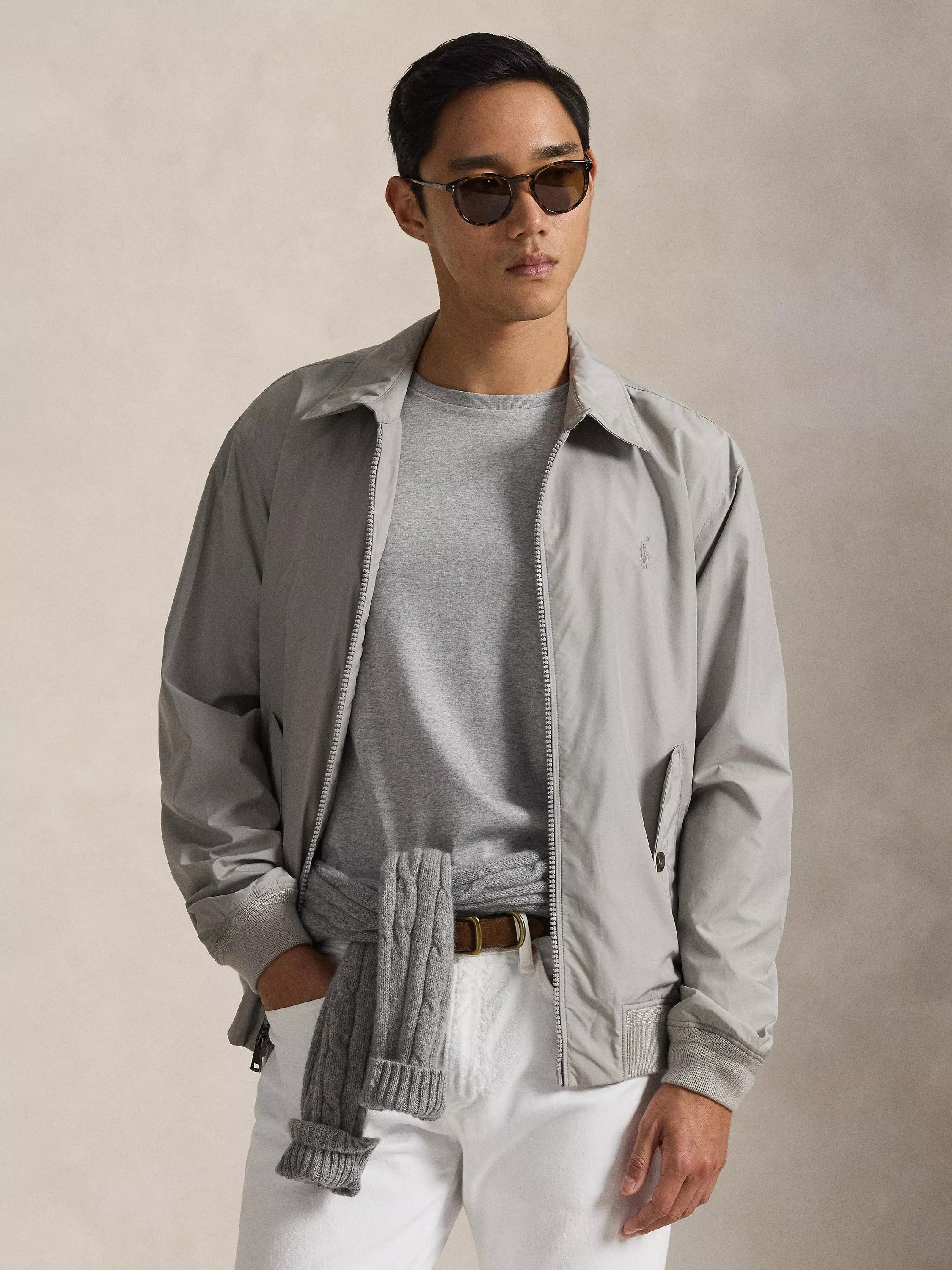 Man in sunglasses wearing a grey jacket over a t-shirt, with a sweater tied at the waist and white trousers.