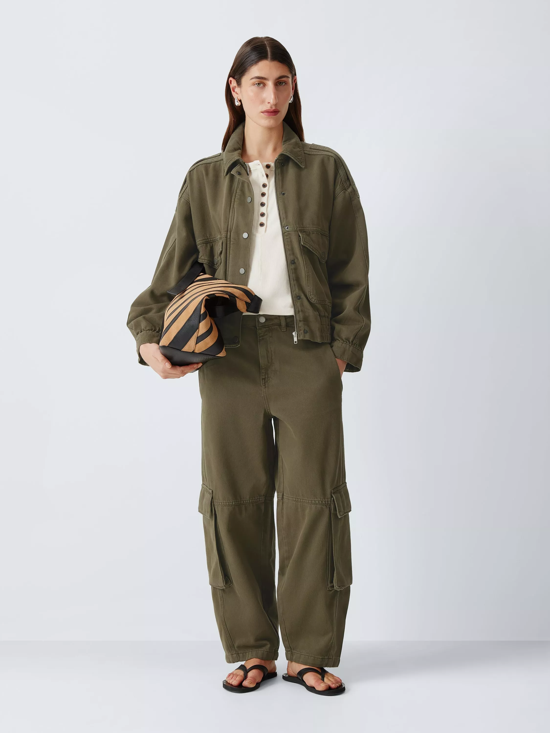 A woman sits on the floor wearing a sleeveless cream top, olive trousers with white trim, and beige woven shoes.