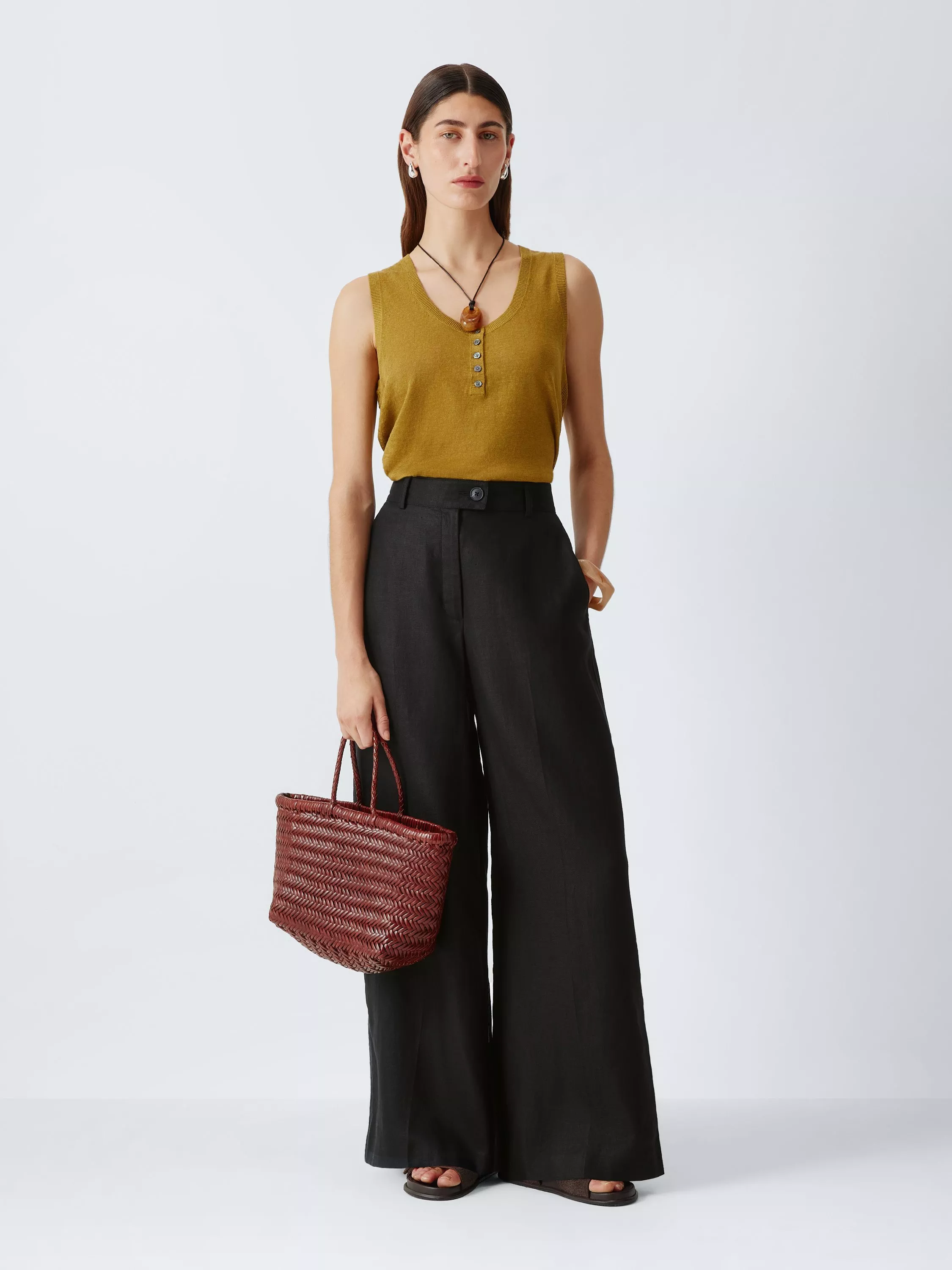 Woman wearing mustard sleeveless knit top with wide-leg black trousers, holding woven tote bag in studio setting