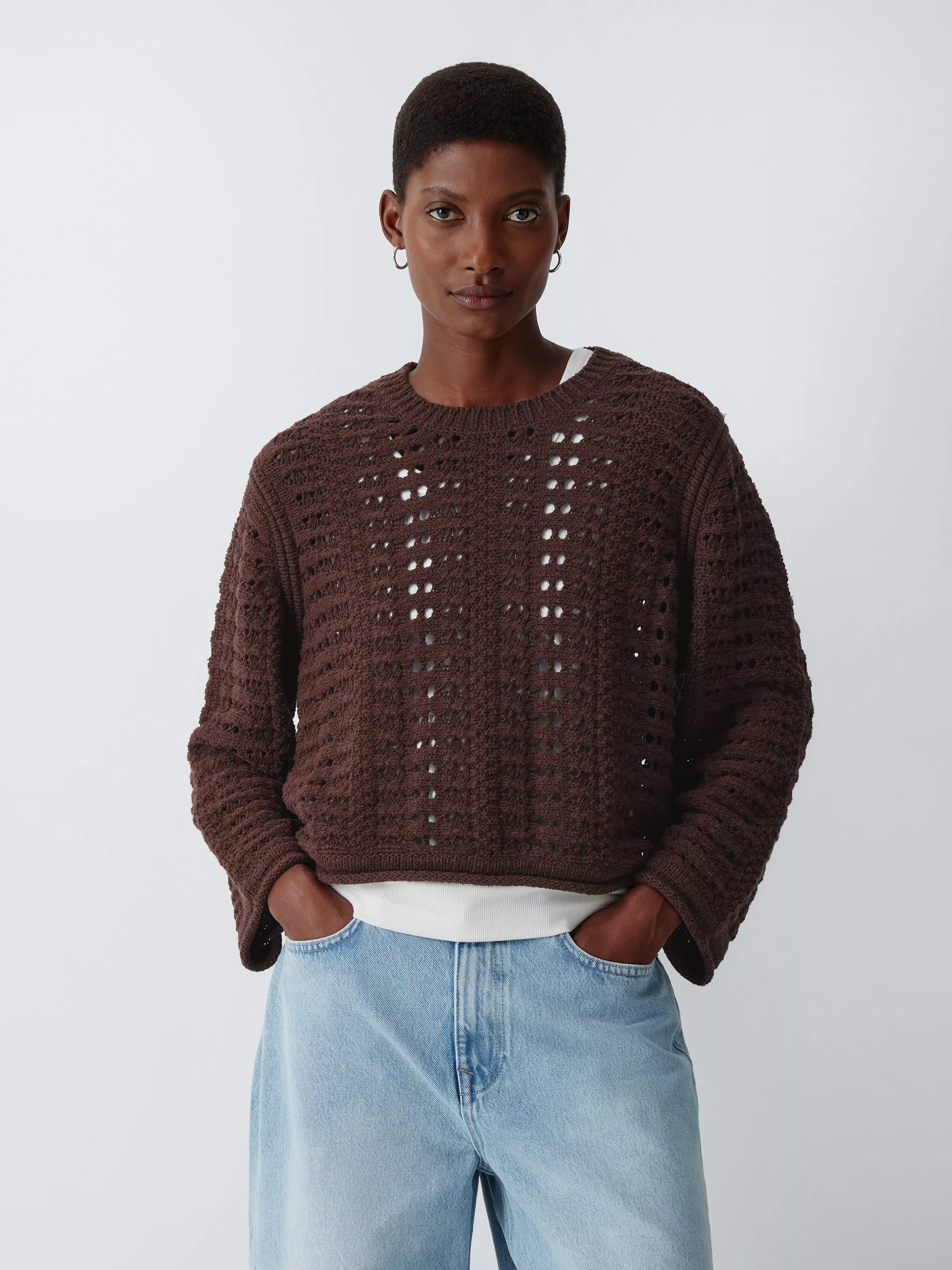 Model wearing brown open-knit jumper with long sleeves and light blue jeans, hands in pockets, studio background
