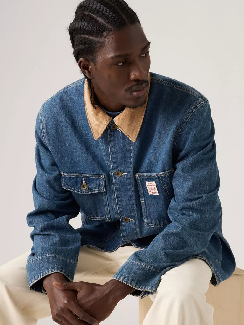 Man wearing blue denim jacket with tan collar and off-white trousers, sitting thoughtfully against a plain background.