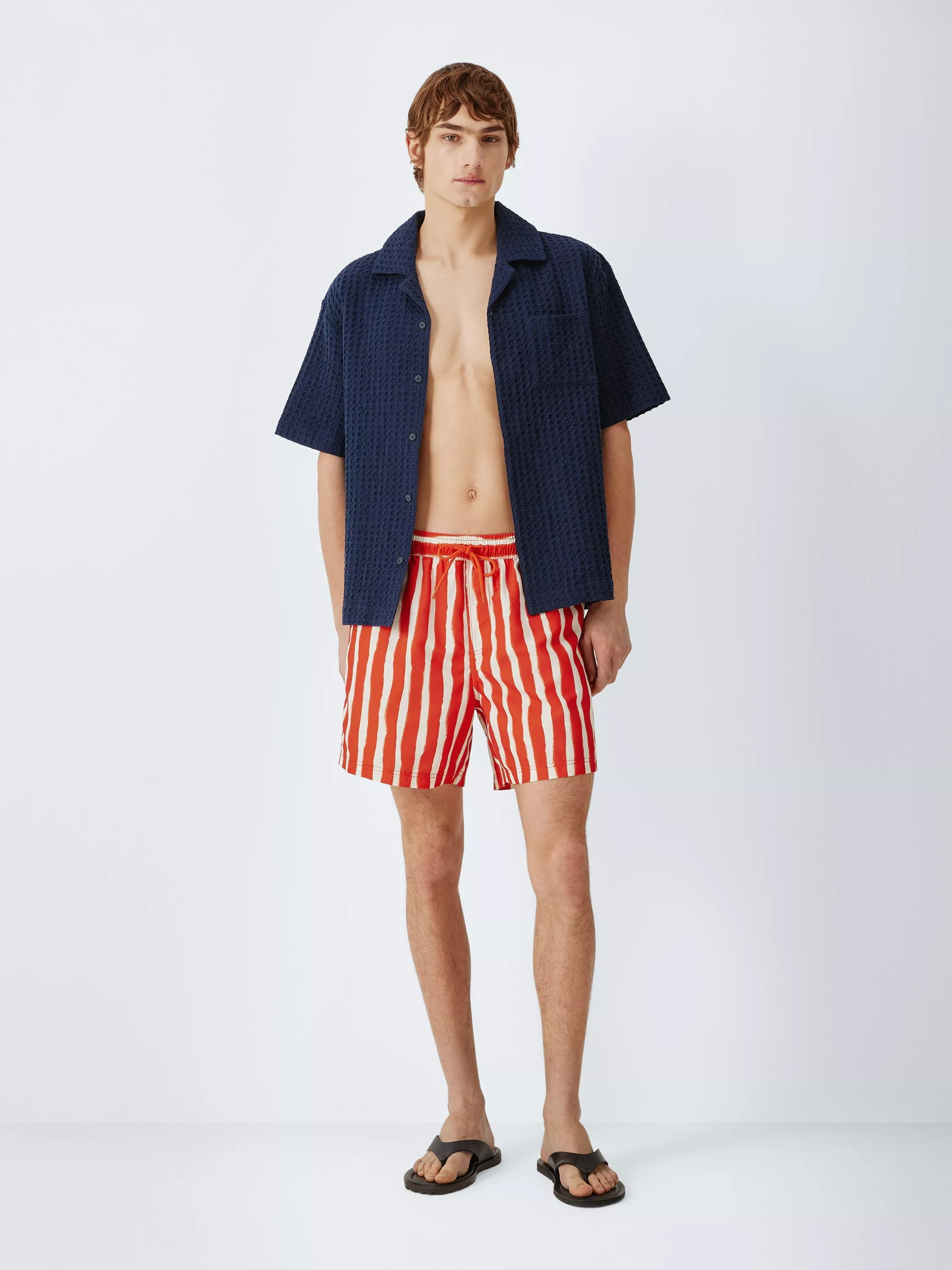 Male model wearing navy textured short-sleeve shirt with red and white striped swim shorts and flip-flops, studio shot