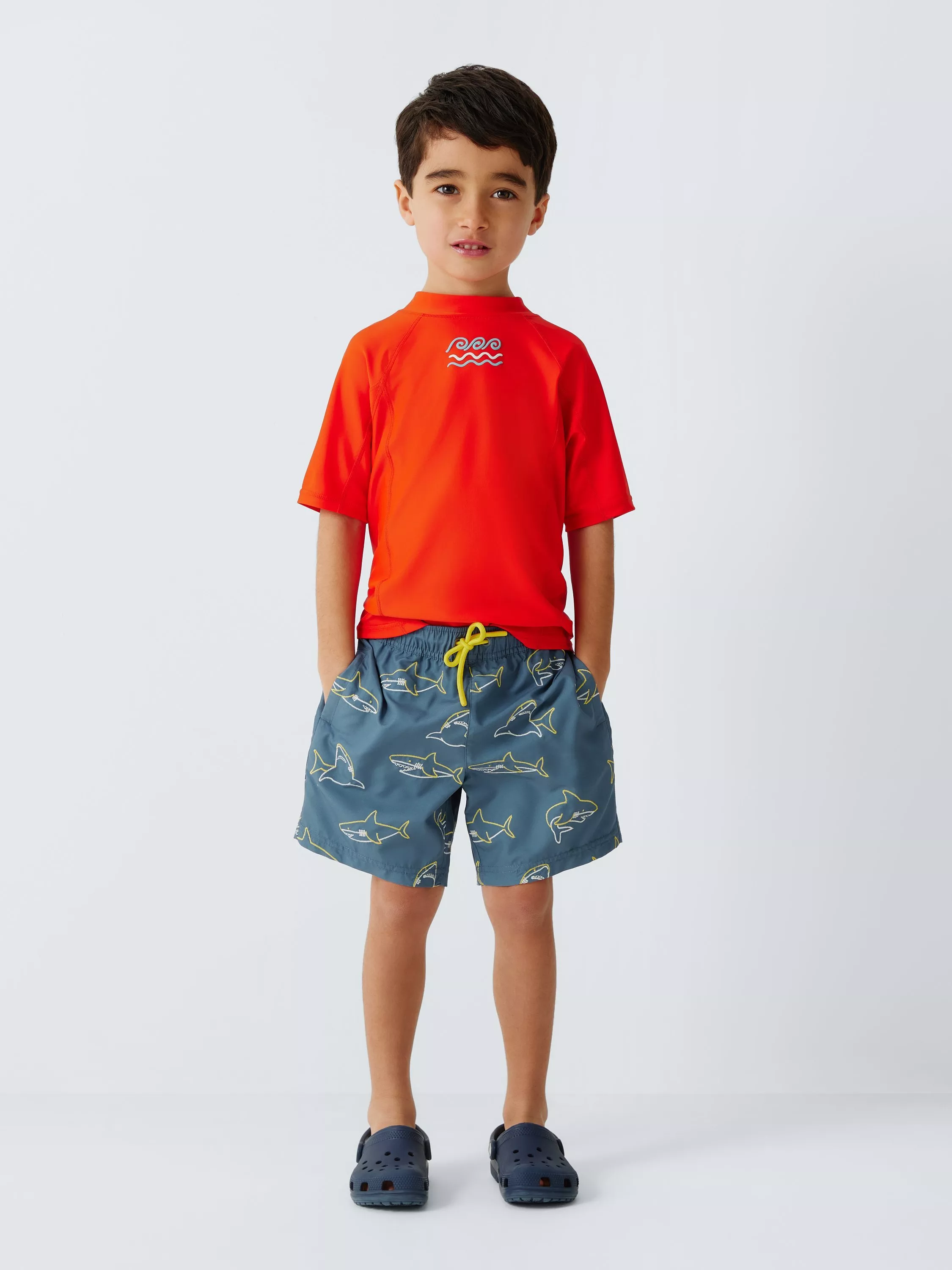 Child wearing red short-sleeve rash vest, blue shark-print swim shorts and navy clogs on grey studio background