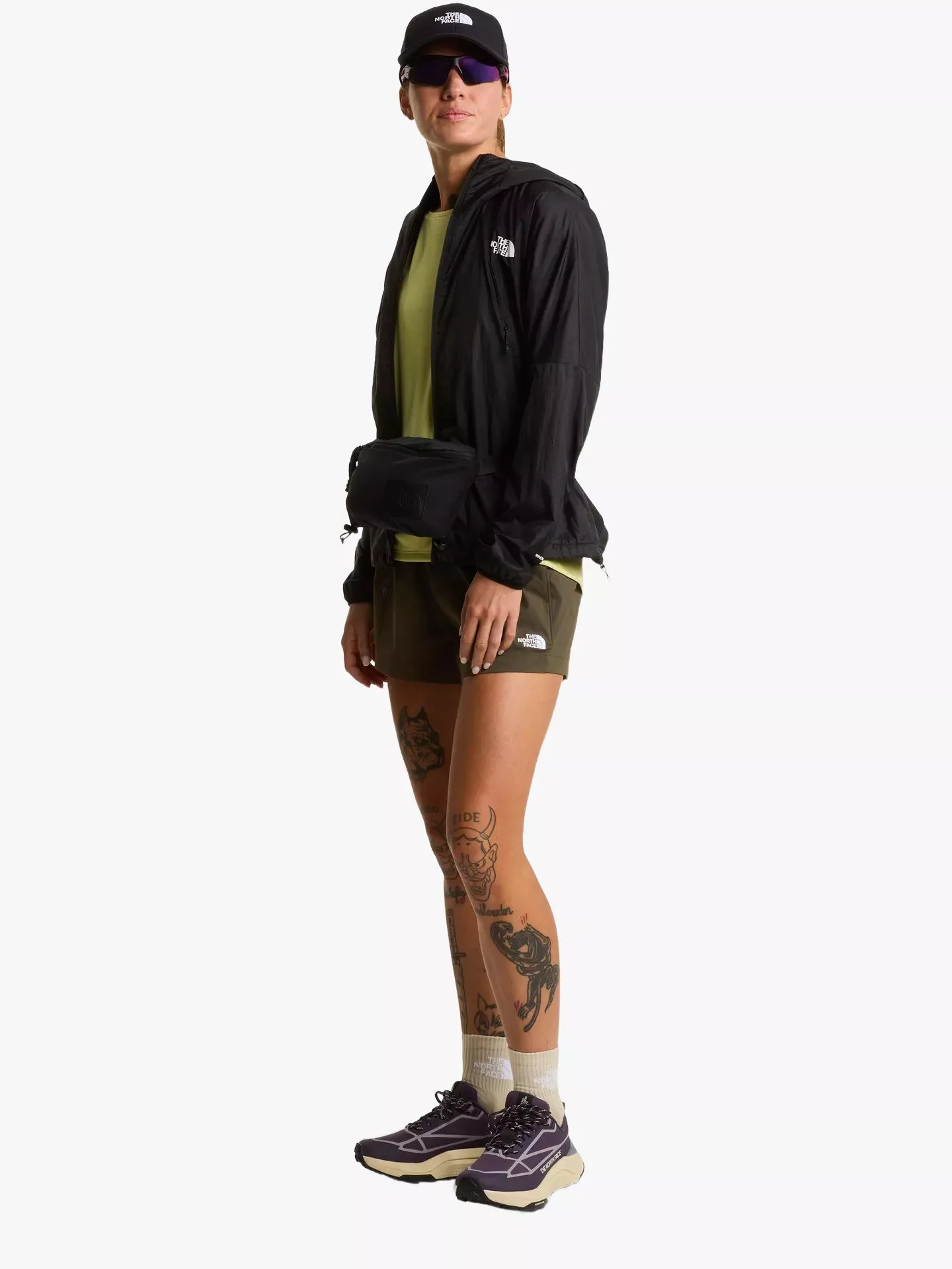 Female hiker in black windbreaker, cap and shorts with waist pack, standing in trail running shoes.