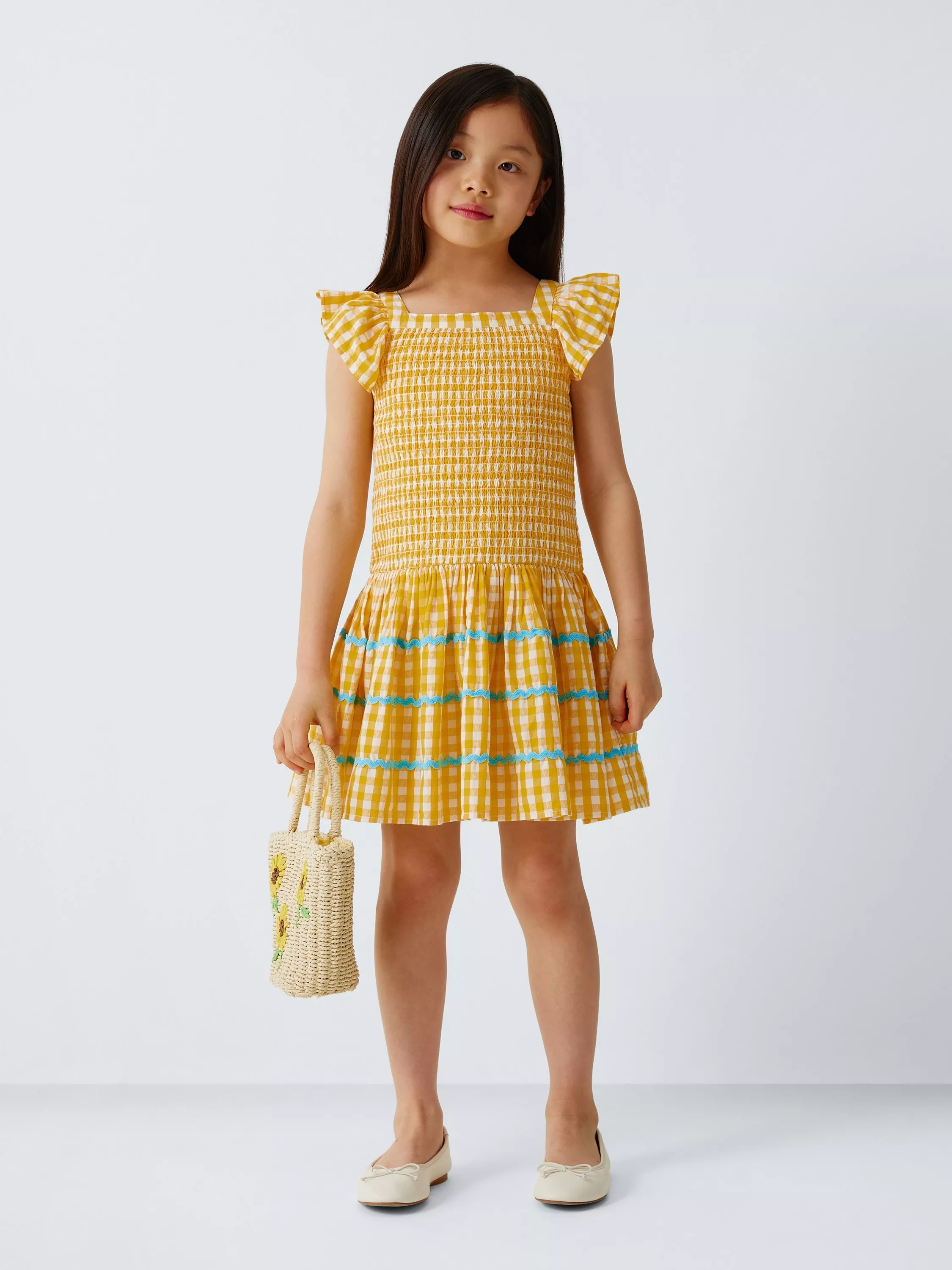 Girl in yellow gingham smocked summer dress with ruffle sleeves, blue trim, holding straw tote bag