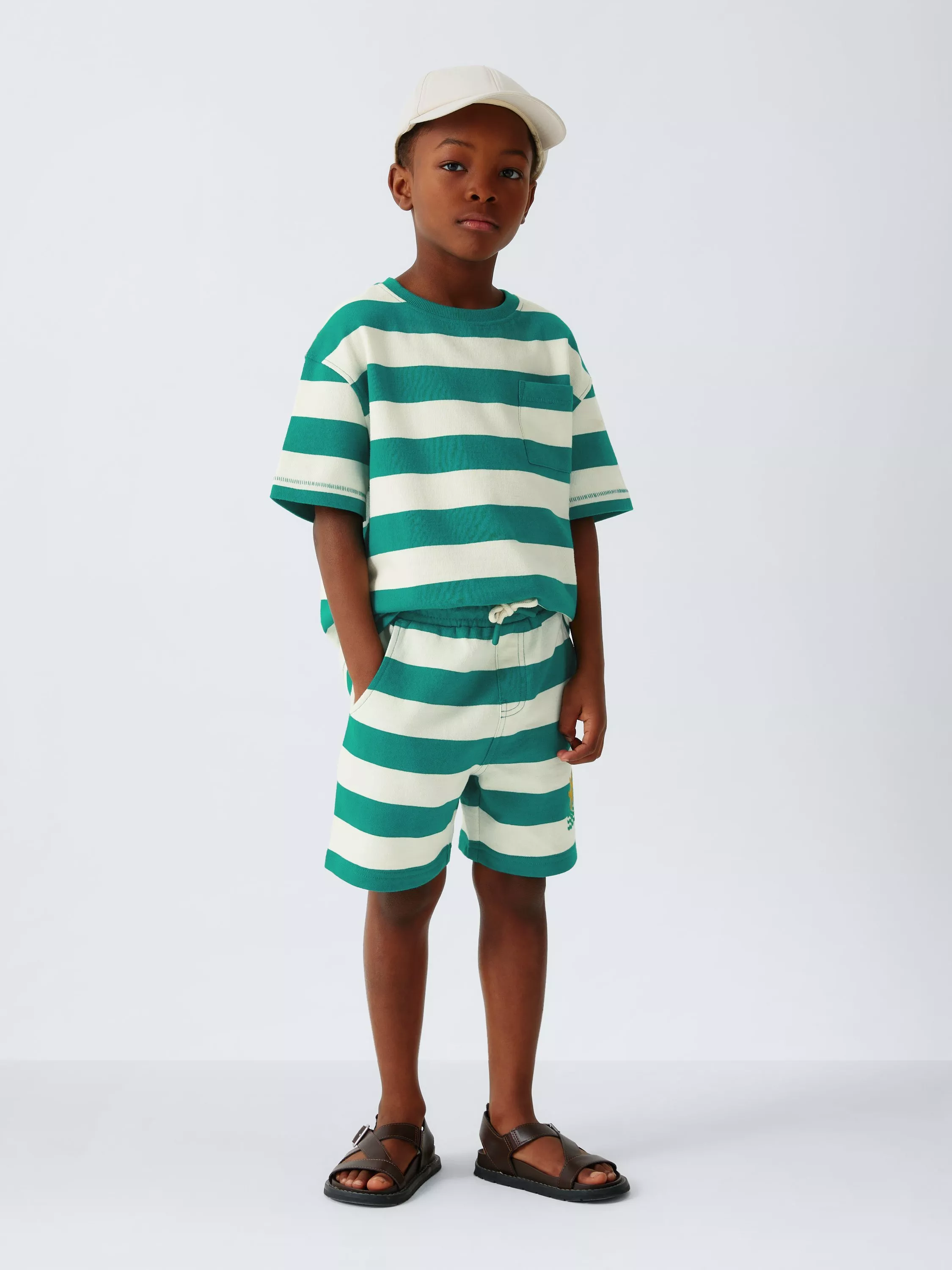 Young boy in green and white striped T-shirt and shorts set, wearing a white cap and brown sandals in studio