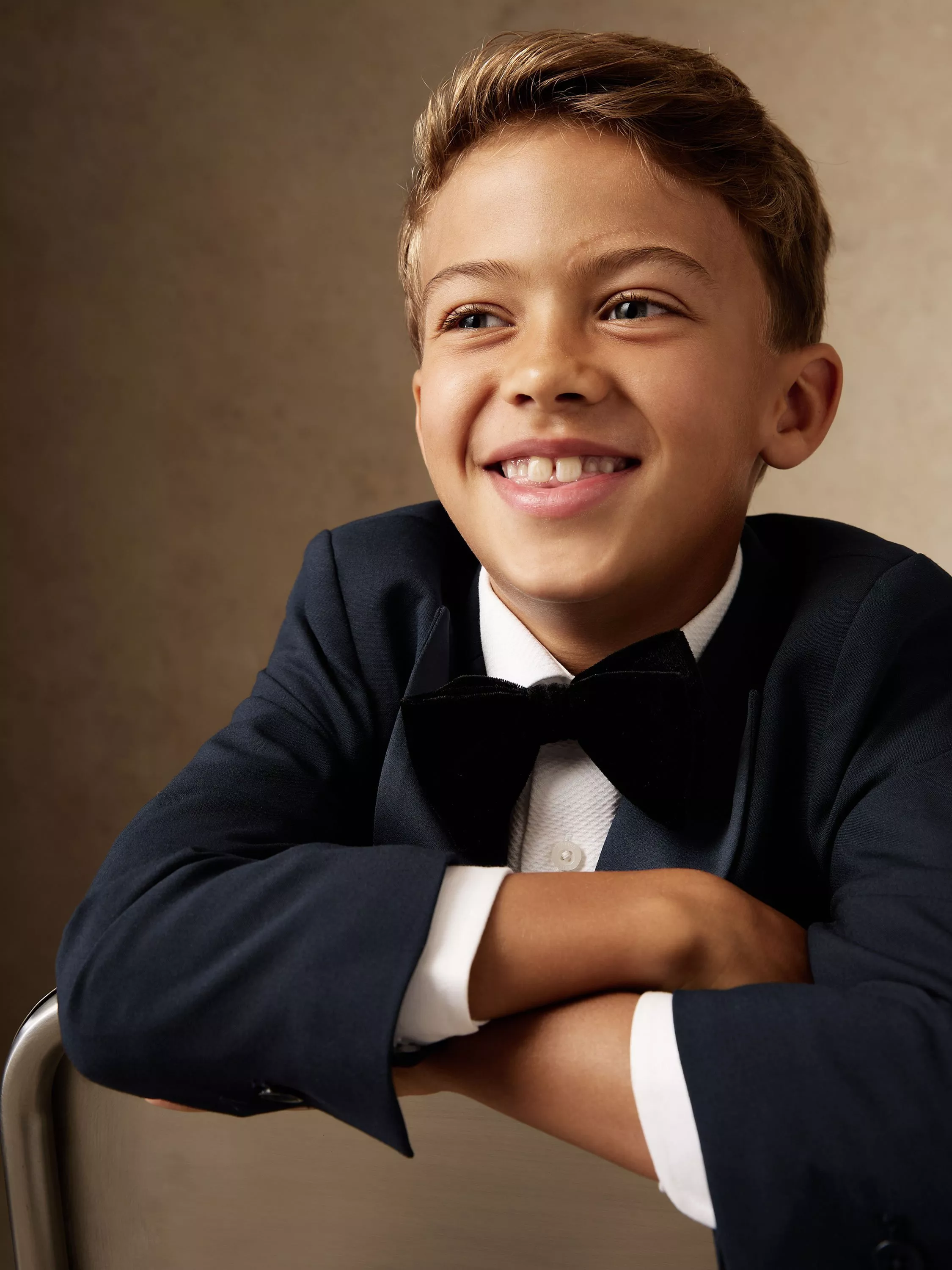 Smartly dressed boy in a dark suit and bow tie, smiling with arms folded, in front of a neutral backdrop.