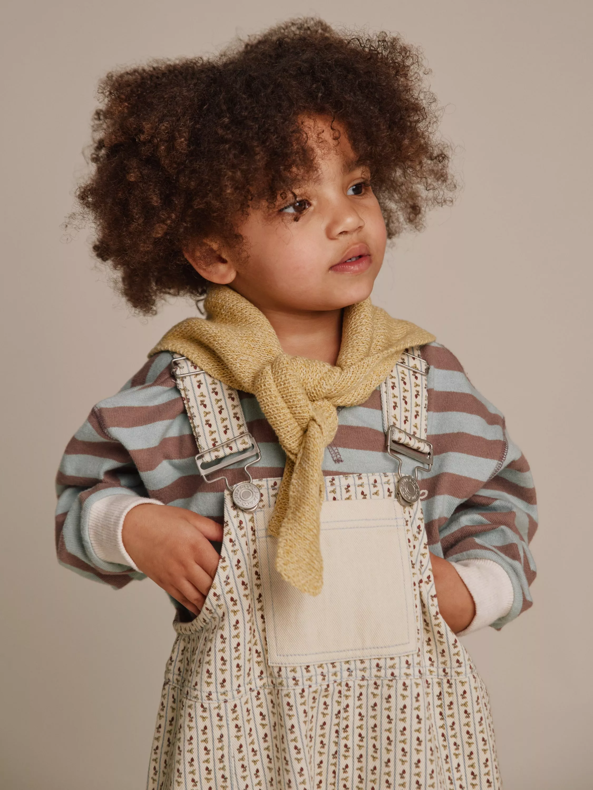 Toddler in striped jumper, floral dungarees and knitted scarf, studio portrait with curly hair on beige background
