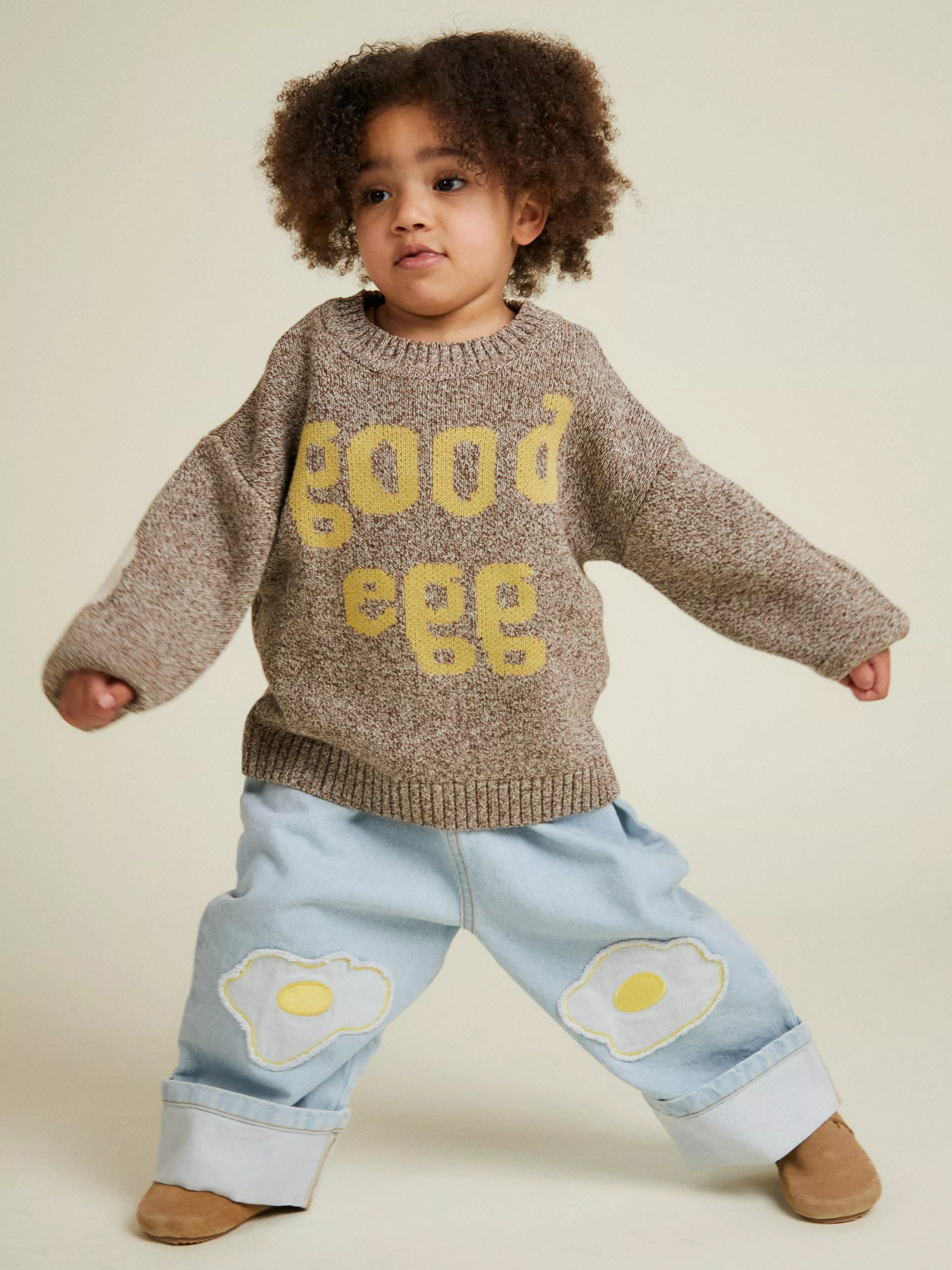 Toddler wearing “good egg” knit jumper and light blue jeans with fried egg patches, standing in studio pose