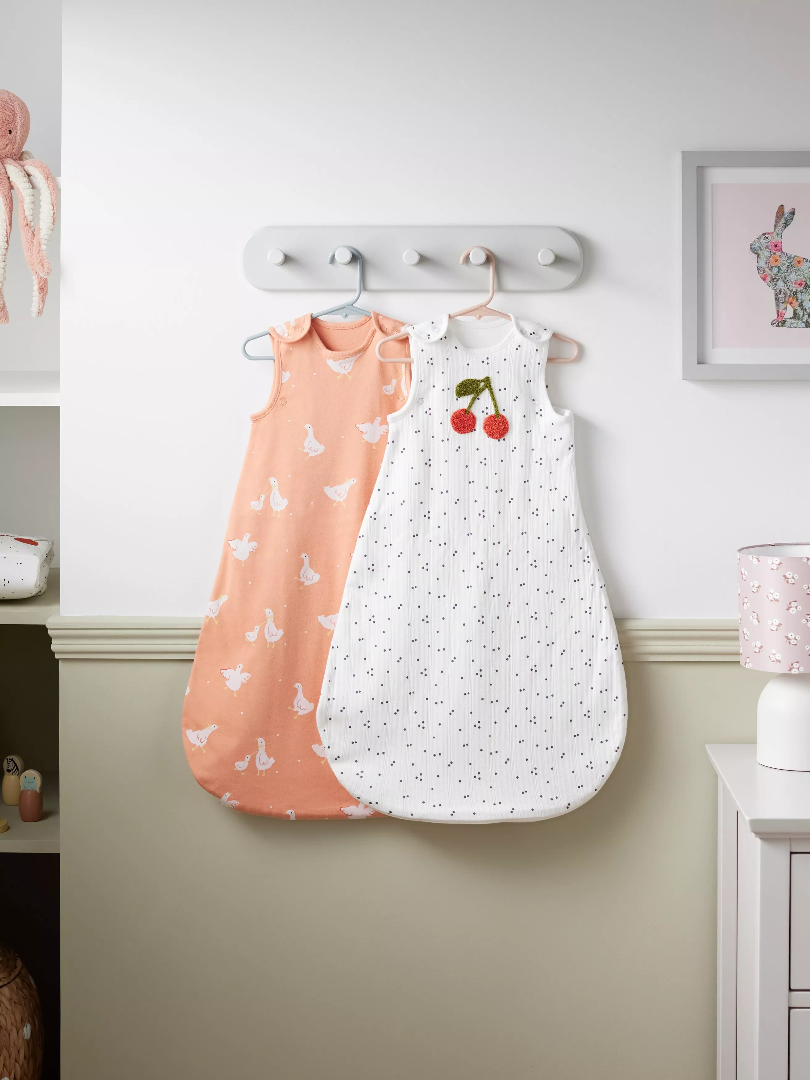 Two baby sleeping bags hanging on nursery wall hooks, one pink goose print and one white with cherry detail