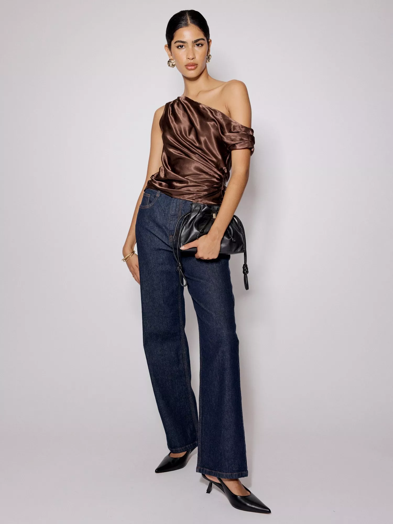 A woman in a brown one-shoulder top and dark jeans, holding a black purse and wearing black heels.