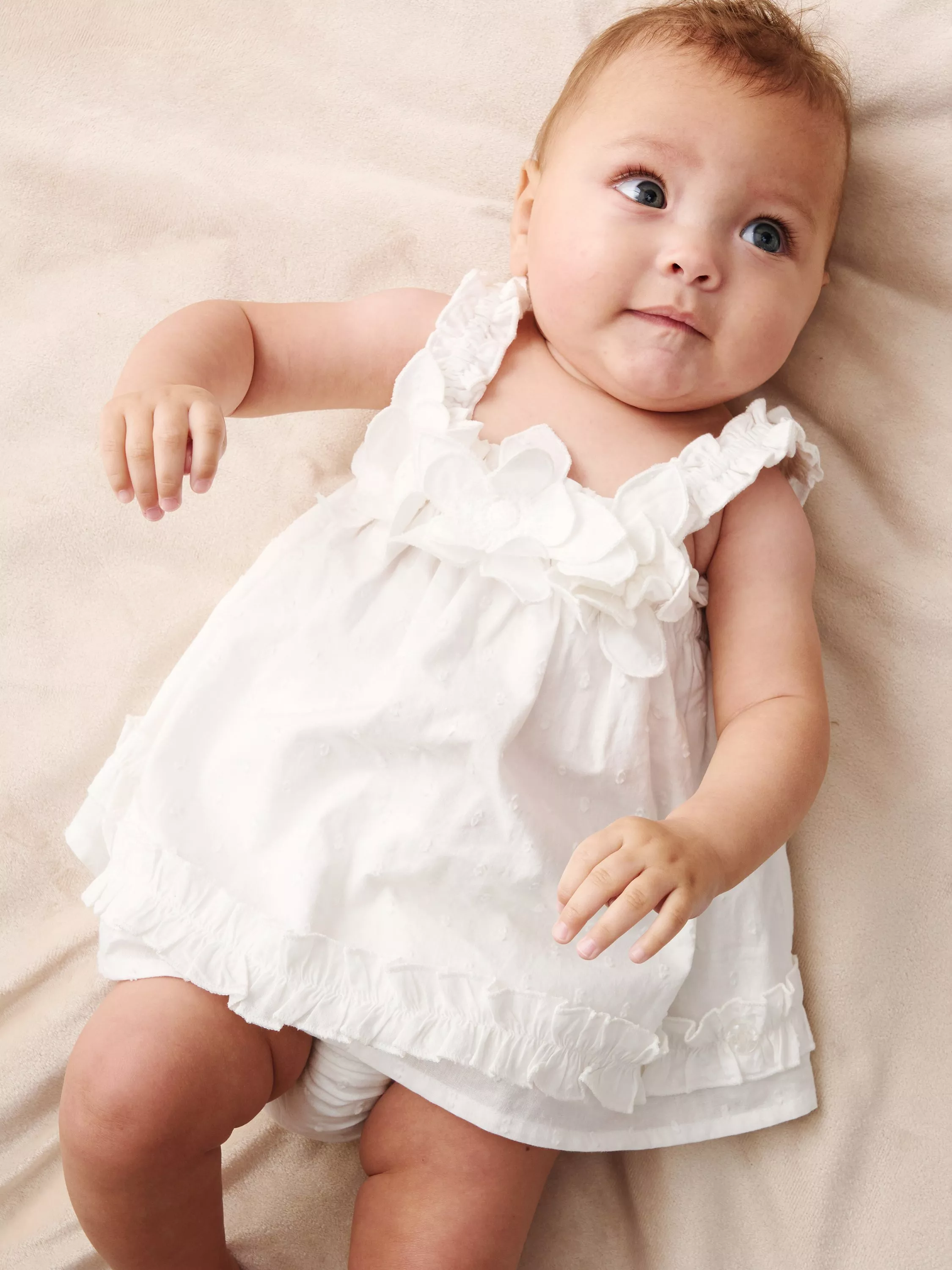A baby in a white dress with floral details lies on a soft beige blanket, looking upwards with a curious expression.