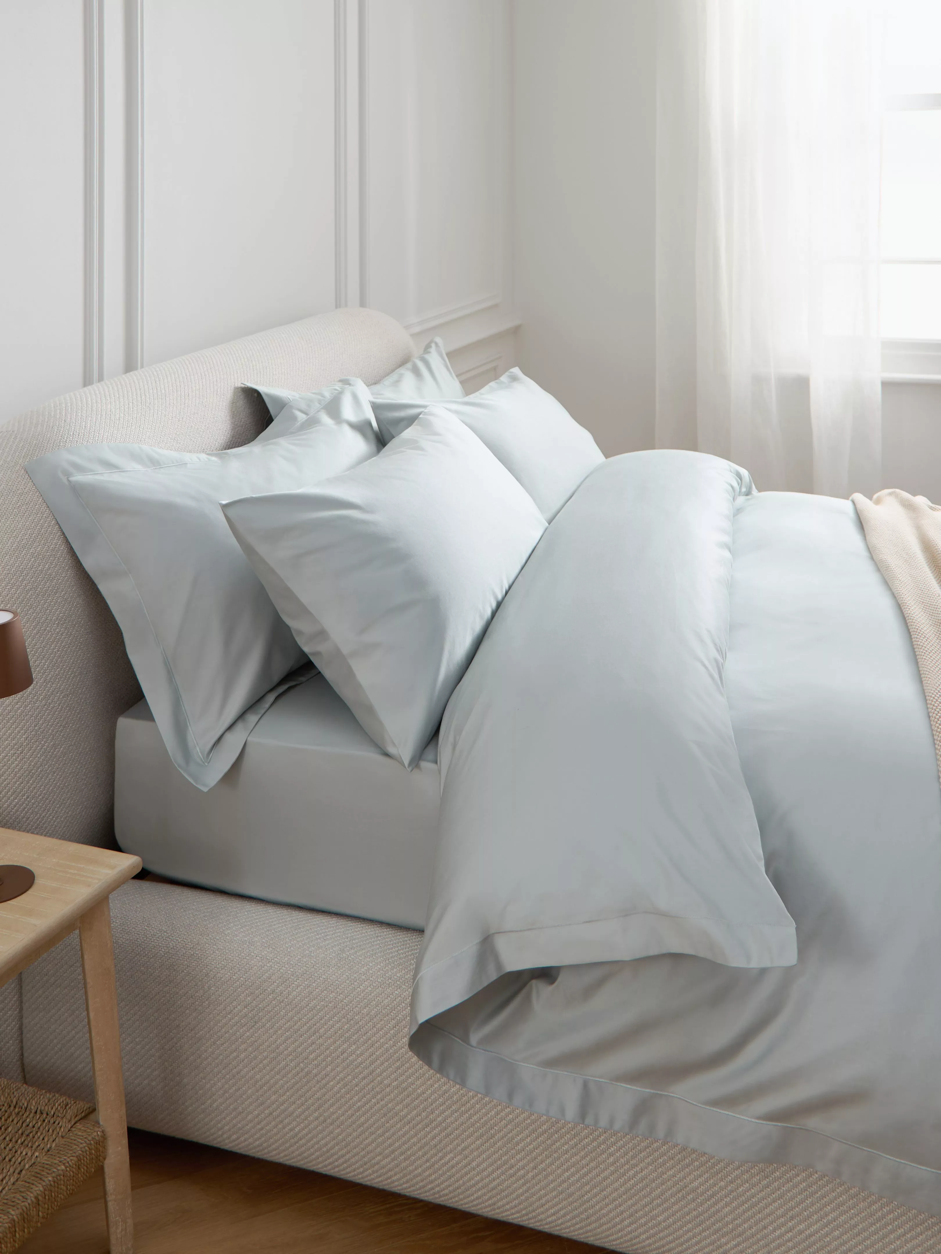 Elegant bedroom with soft grey sheets and pillows on a plush bed. Sunlight filters through sheer curtains.