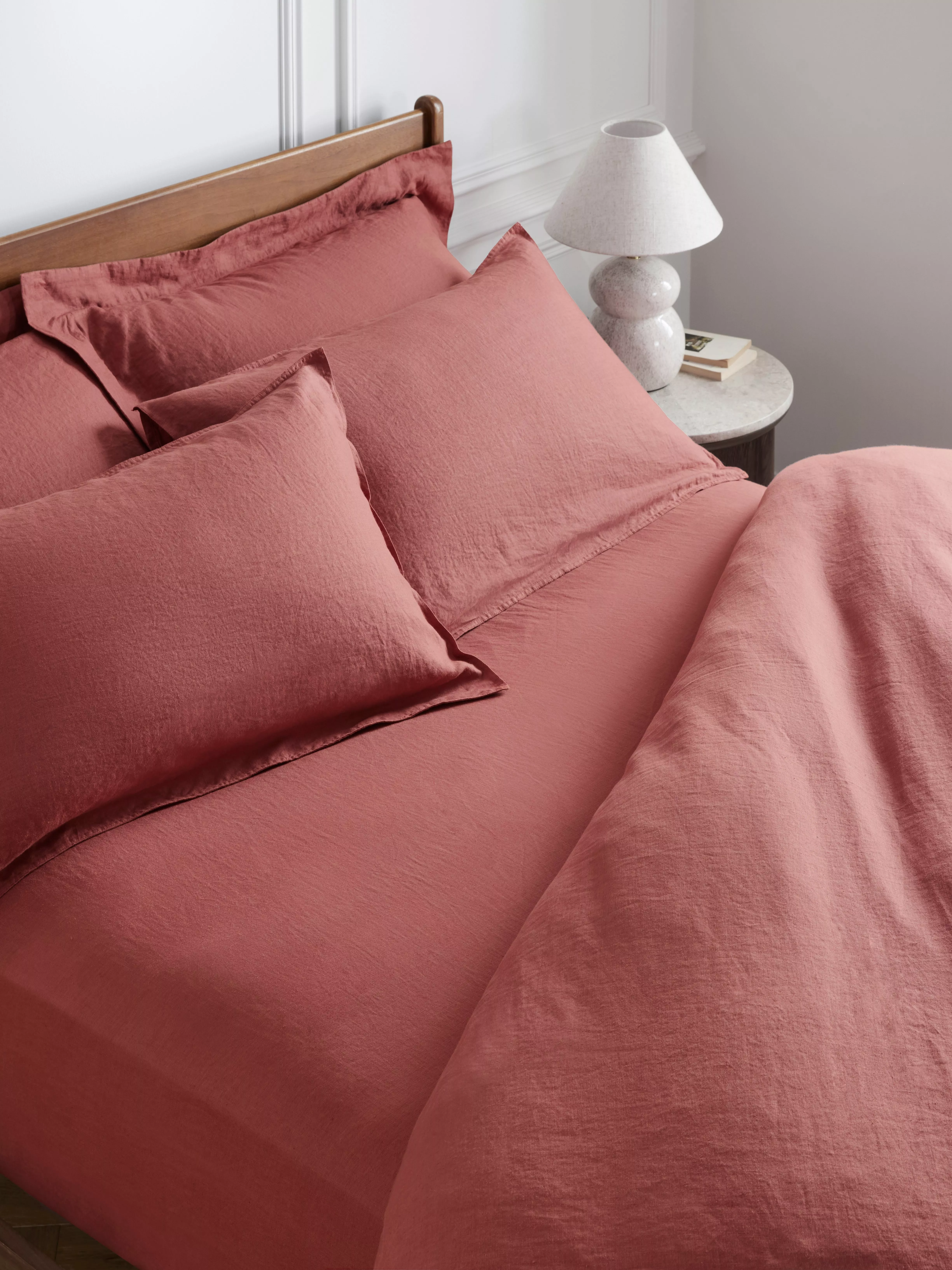 Pink linen bed set with pillows and duvet on a wooden bed frame, next to a marble bedside table and lamp.