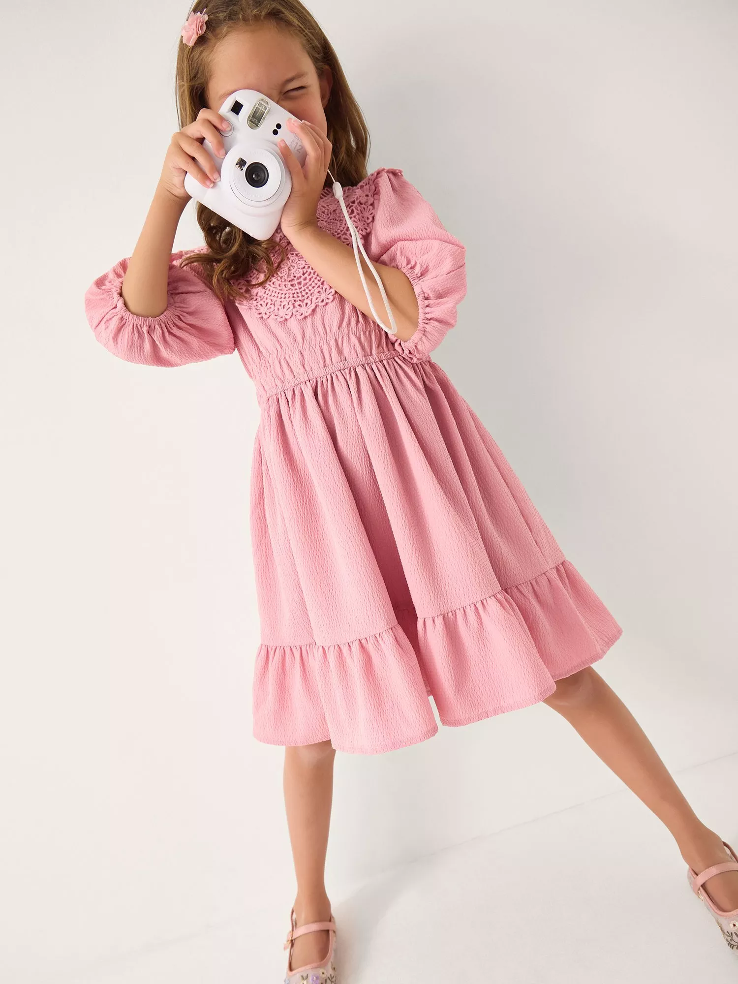 A young girl in a pink dress takes a photo with a white instant camera, smiling in a playful pose.