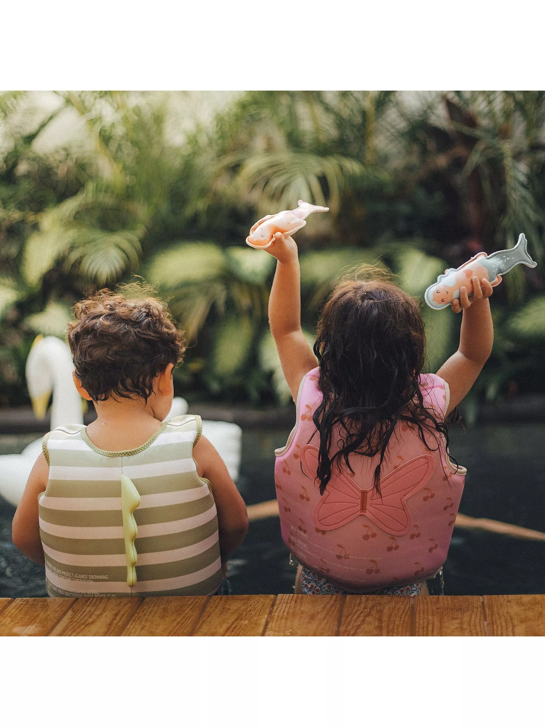 Two children in float vests by a garden pool, holding toy fish, ready for a safe swim outdoors