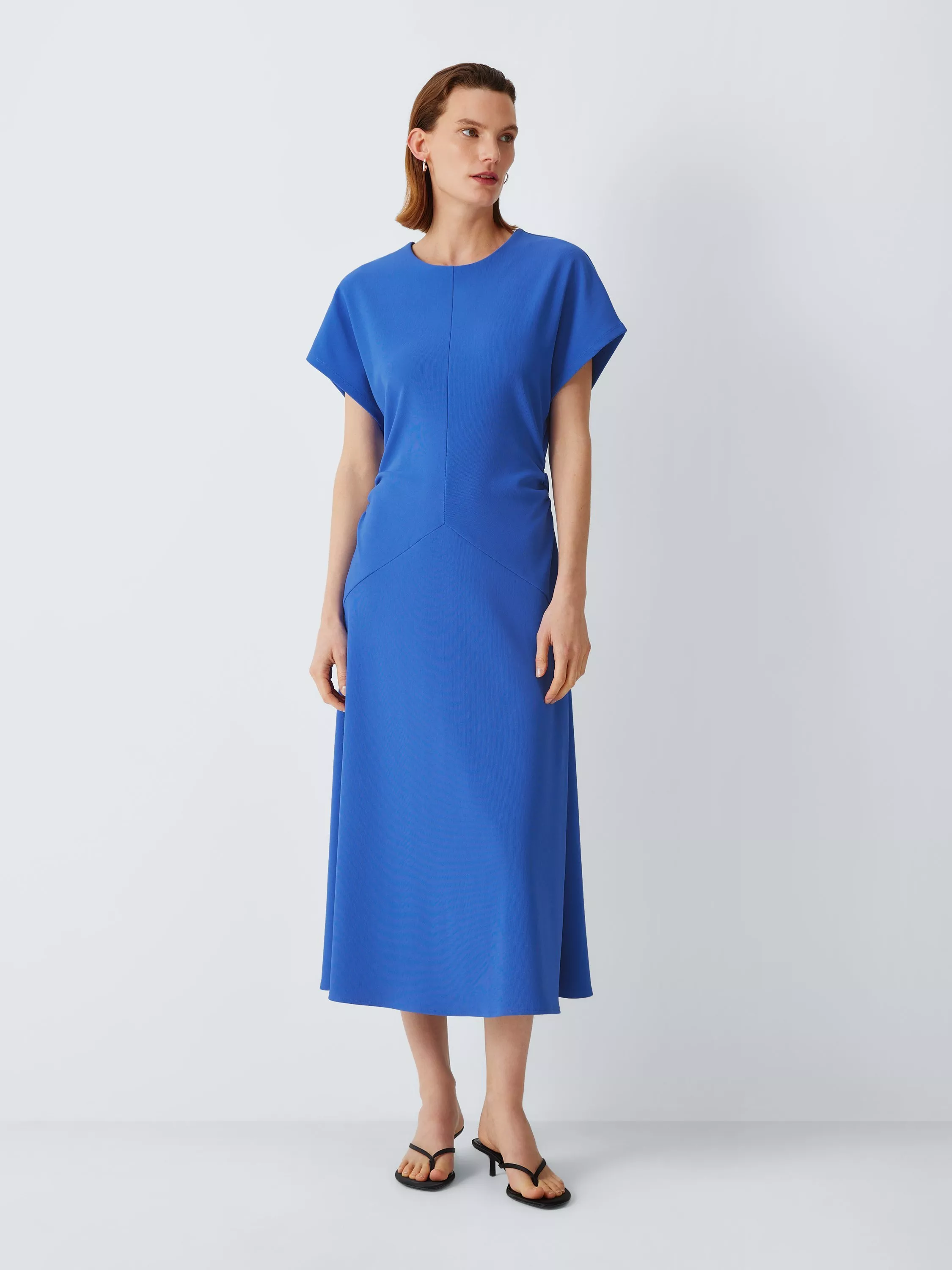 Model wearing a cobalt blue midi dress with cap sleeves and seam detail, styled with black sandals in studio.