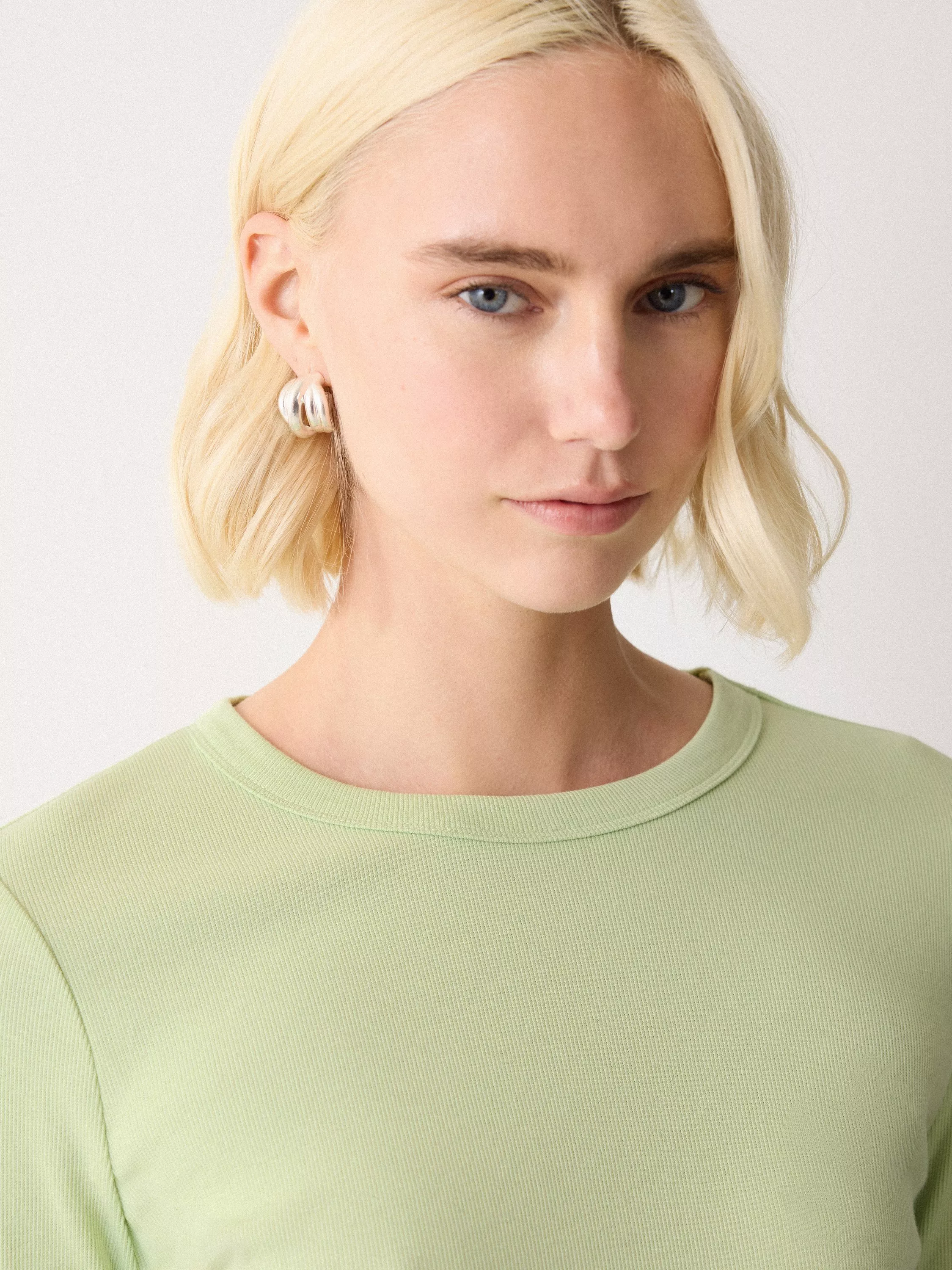 Woman with blonde bob hairstyle, wearing silver hoop earrings and a light green top, facing forward.