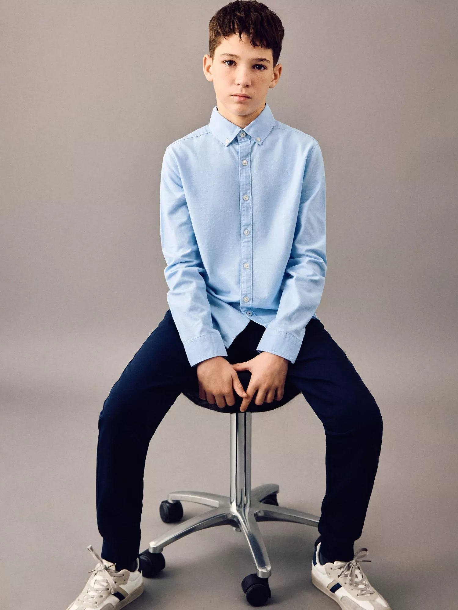 A young boy in a light blue shirt and navy trousers sits on a stool against a grey background, wearing trainers.