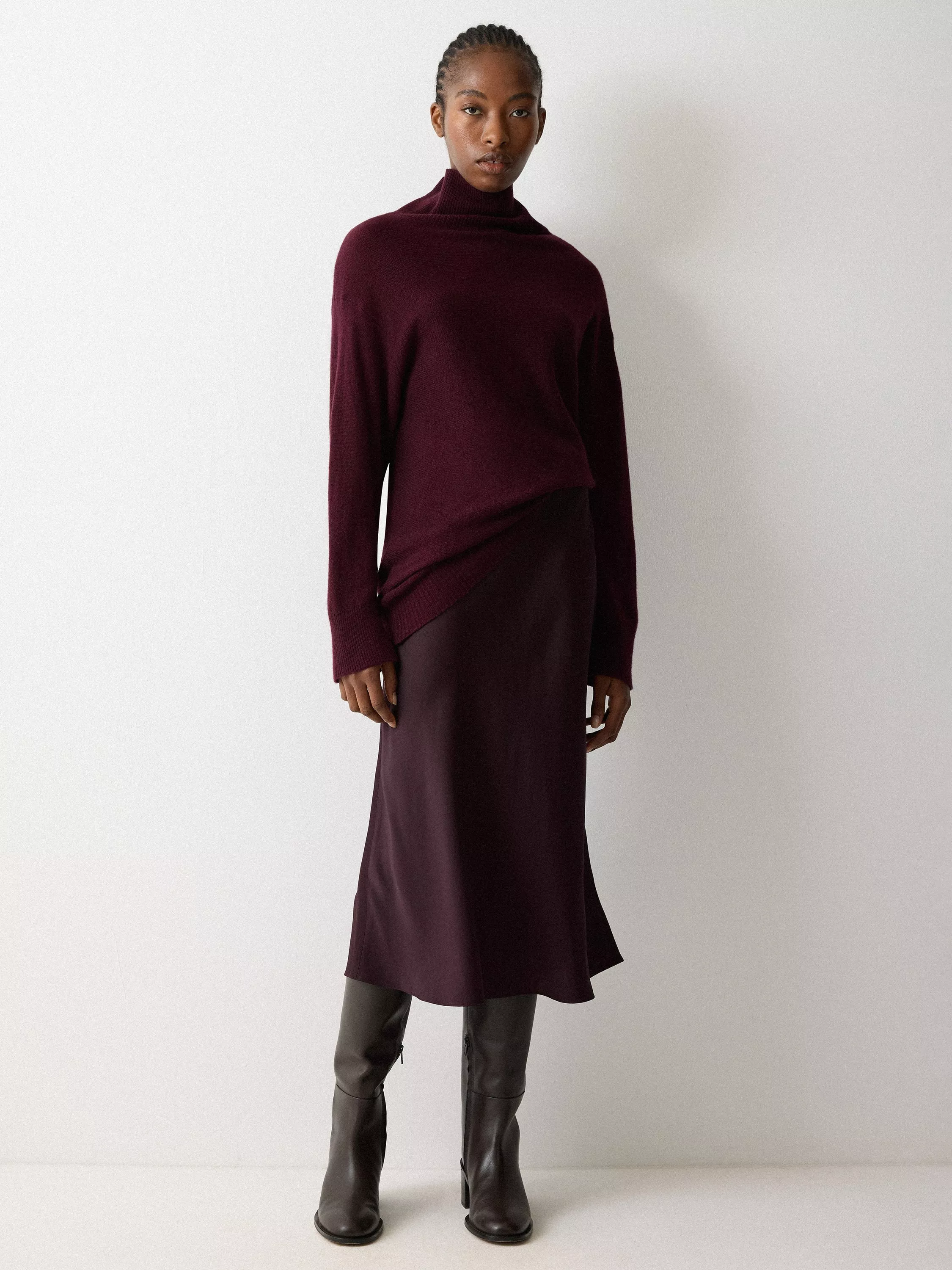 A person wearing a burgundy sweater and skirt paired with dark knee-high boots, stands against a light wall.