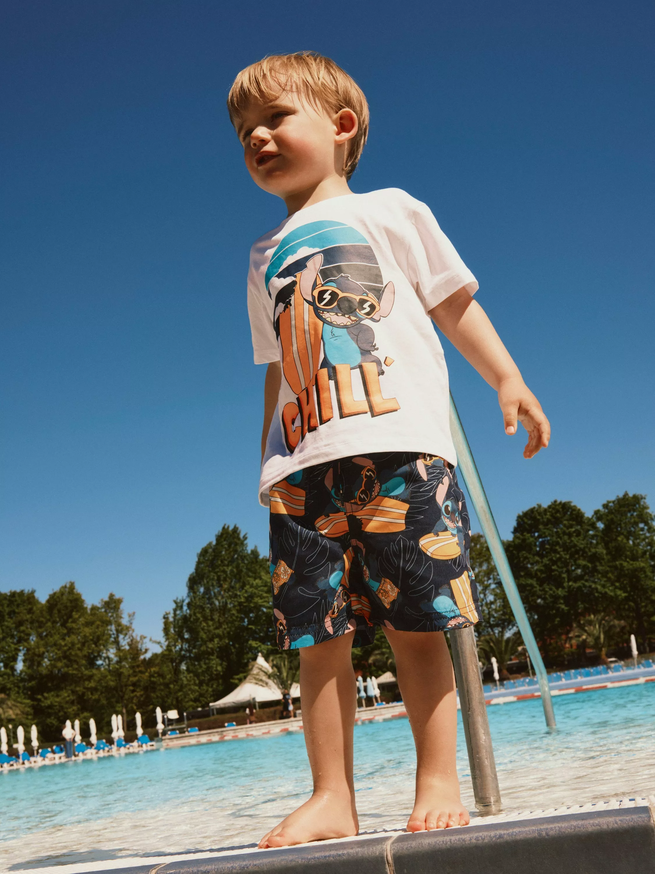 Young boy in Disney Stitch T-shirt and patterned swim shorts standing by outdoor pool on sunny day